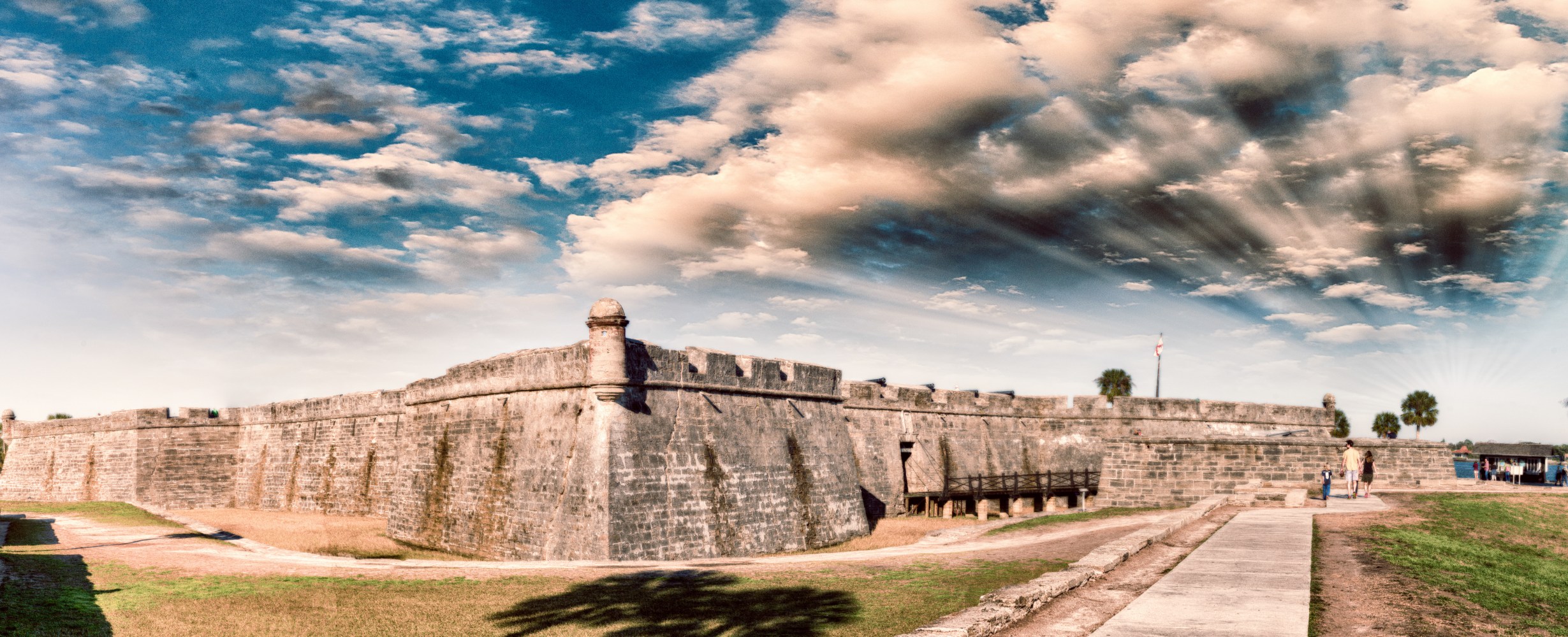 Historical stone fort under a partly cloudy sky with sun rays, path leading to entrance.