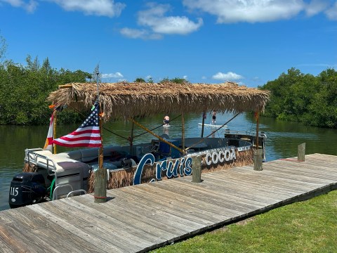 Tiki boat with flags at a dock under a sunny sky, surrounded by greenery.