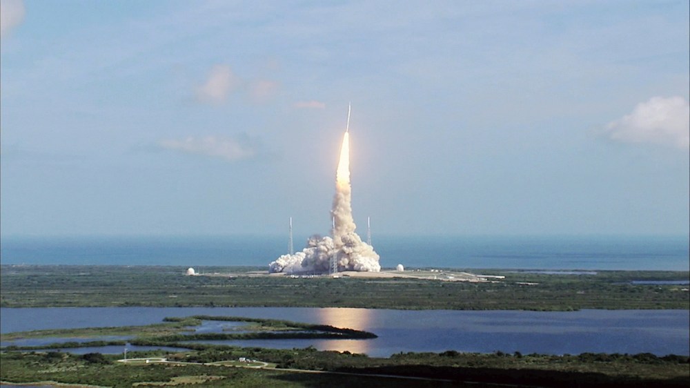 Rocket launching from a pad with smoke plume against a blue sky and water in the foreground.