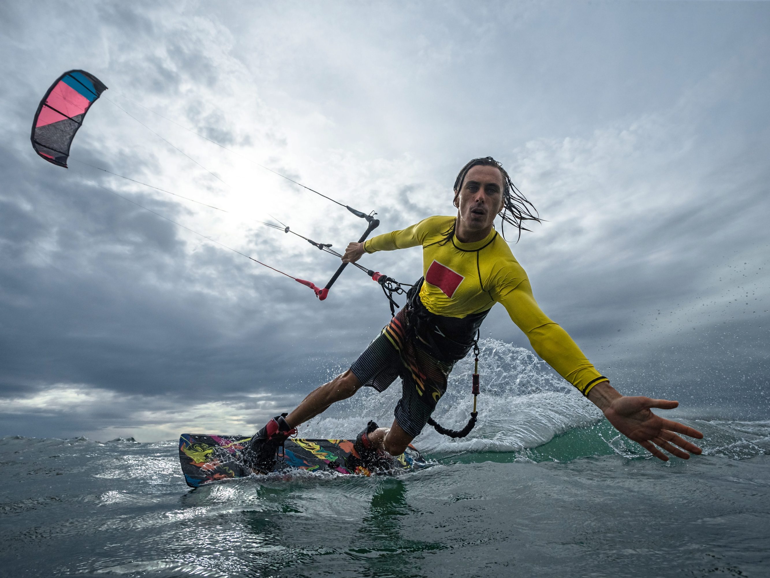 Person kite surfing on wavy ocean under cloudy sky, wearing a yellow wetsuit.