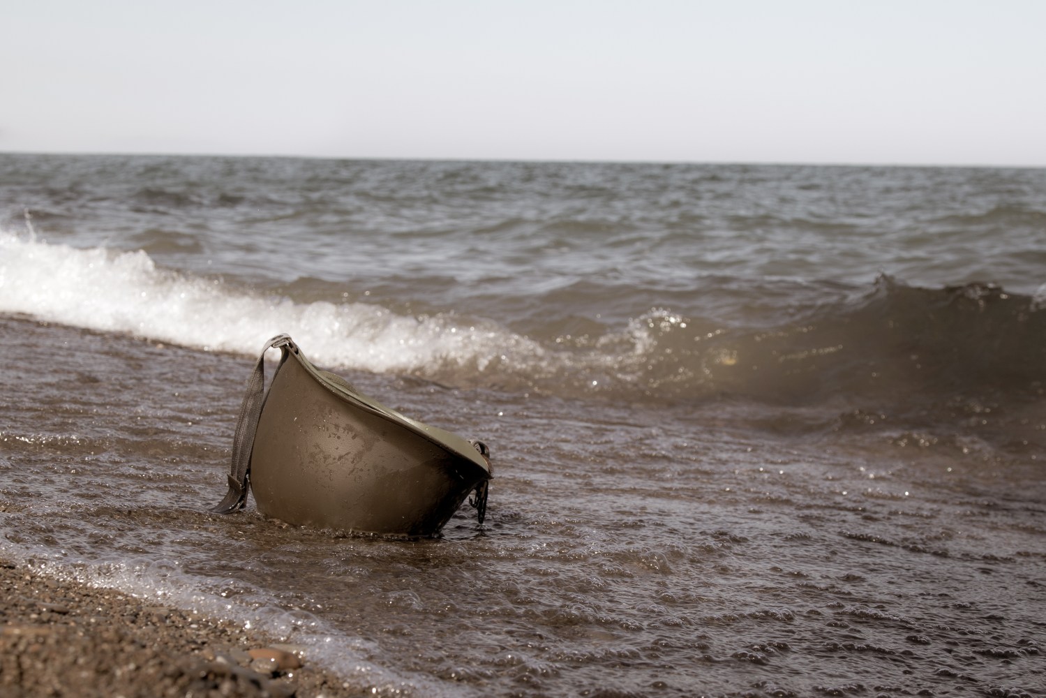 Army helmet partially submerged in ocean waves on a sandy beach.