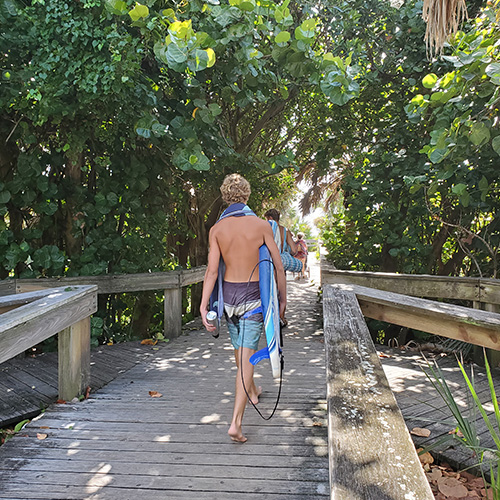 Person in swim trunks walking on a wooden path with a surfboard, surrounded by lush greenery.