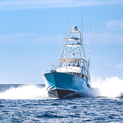 Blue fishing boat cruising on the ocean, creating white waves under a clear sky.