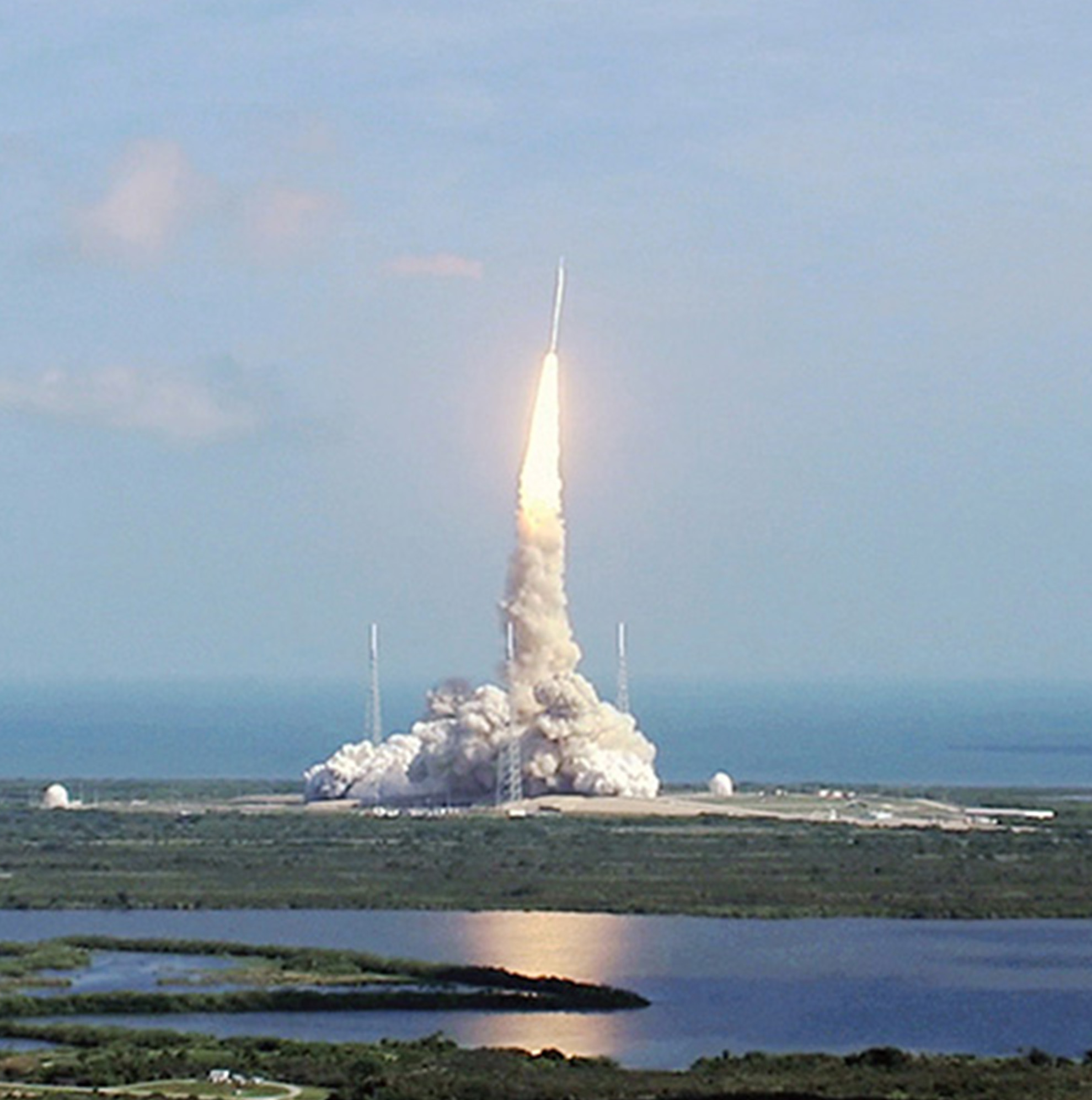 Rocket launching from a pad by the sea, with a trail of smoke, under a clear sky.