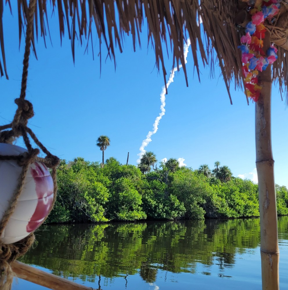 Palm trees by a river, straw roof, and a rocket's smoke trail in a clear blue sky.