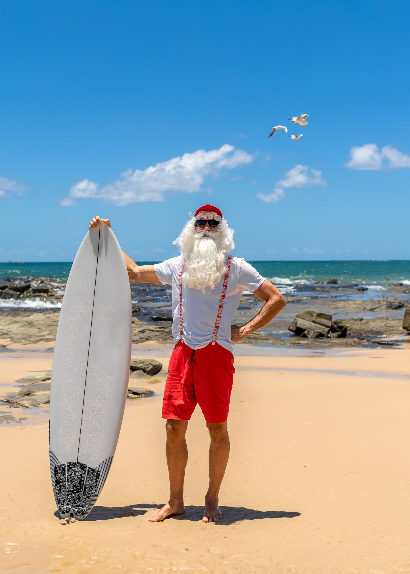 Man dressed as Santa holding a surfboard on a sunny beach with seagulls flying in the background.