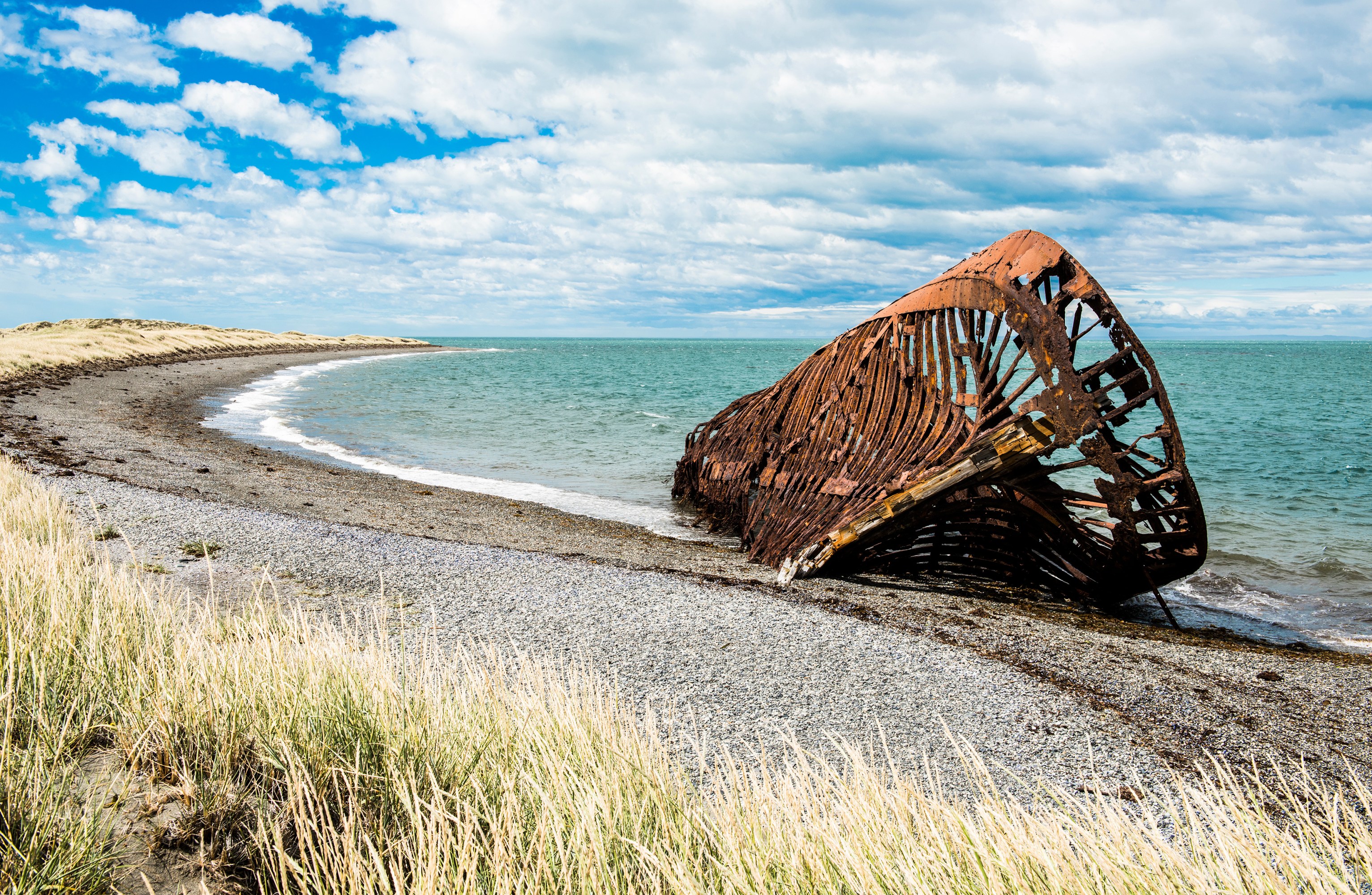 Rusted shipwreck on a rocky beach with grassy dunes and cloudy sky.