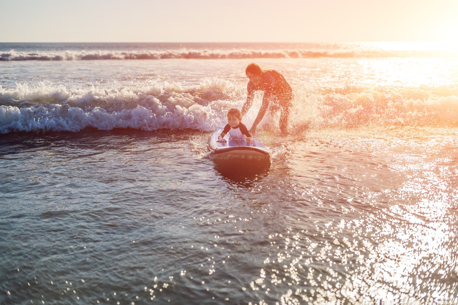 Adult pushing child on surfboard in ocean waves at sunset.