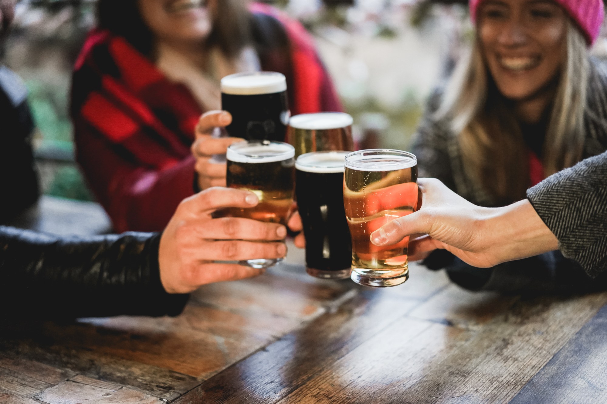 Hands holding beer glasses for a toast around a wooden table, people smiling in background.