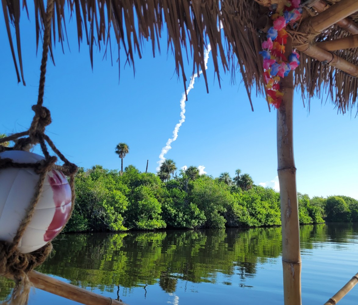 Rocket trail over trees and water, viewed from under a thatched roof.
