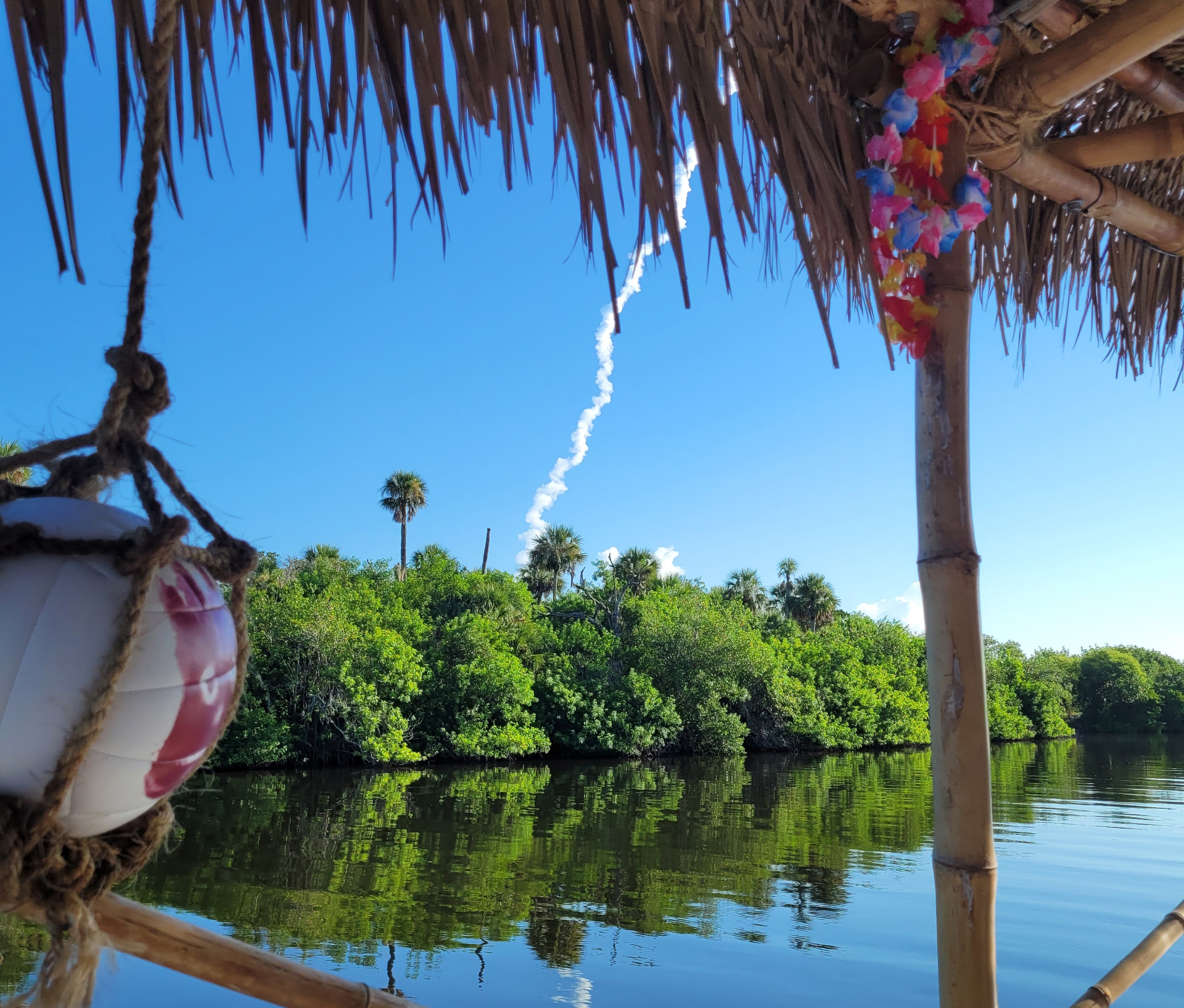Rocket trail over trees and water, viewed from under a thatched roof.