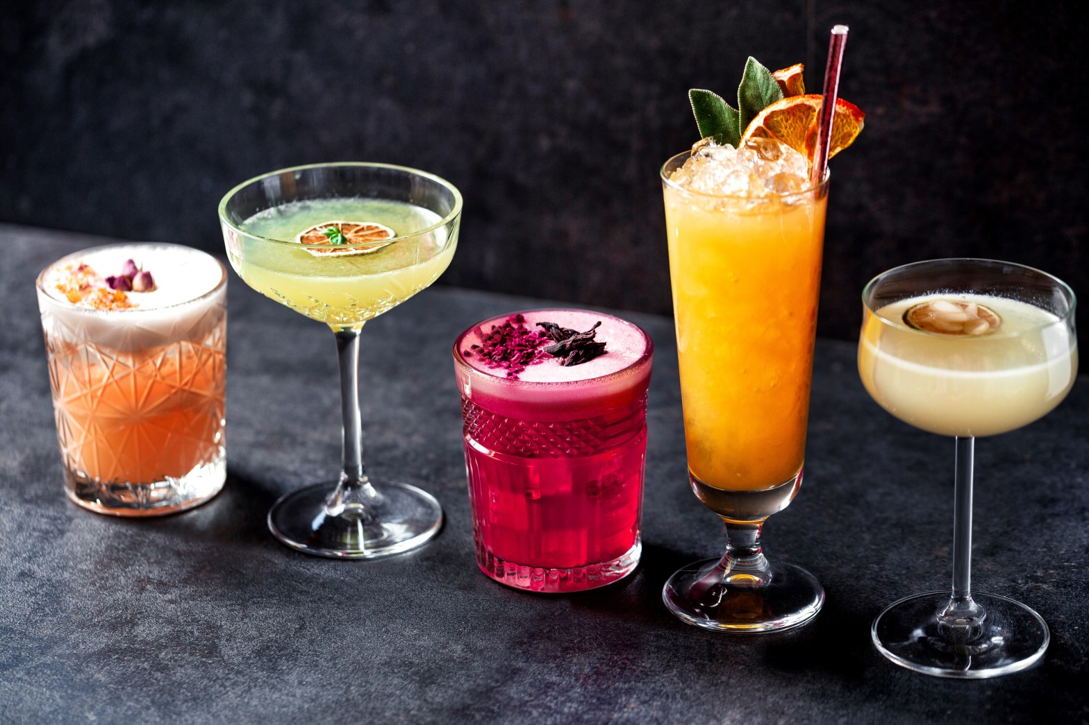 Five colorful cocktails in various glasses on a dark surface.