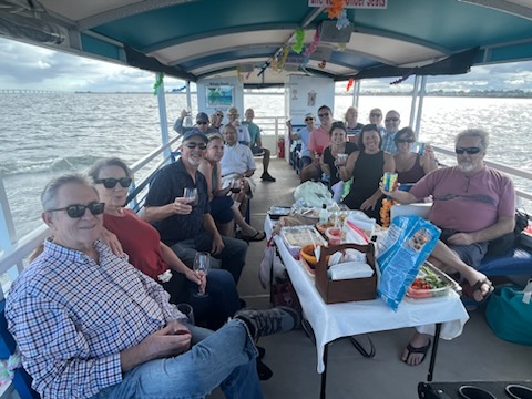 Group of people enjoying a meal on a covered boat, with water visible in the background.