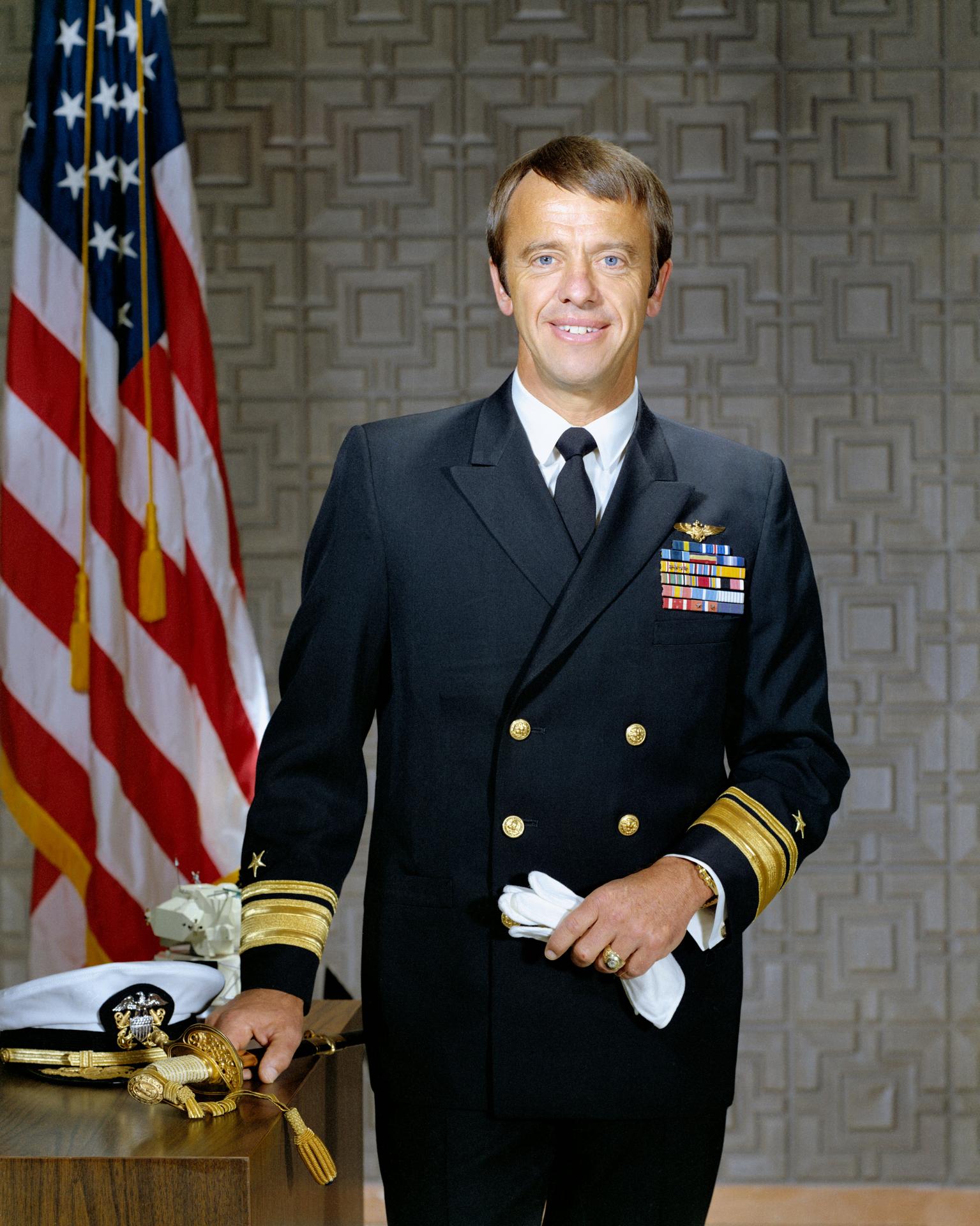 Man in navy uniform with medals, standing by a U.S. flag and a sword on a table.