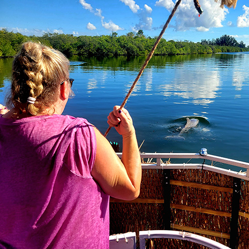 Woman on boat feeds dolphin in calm lake surrounded by trees.