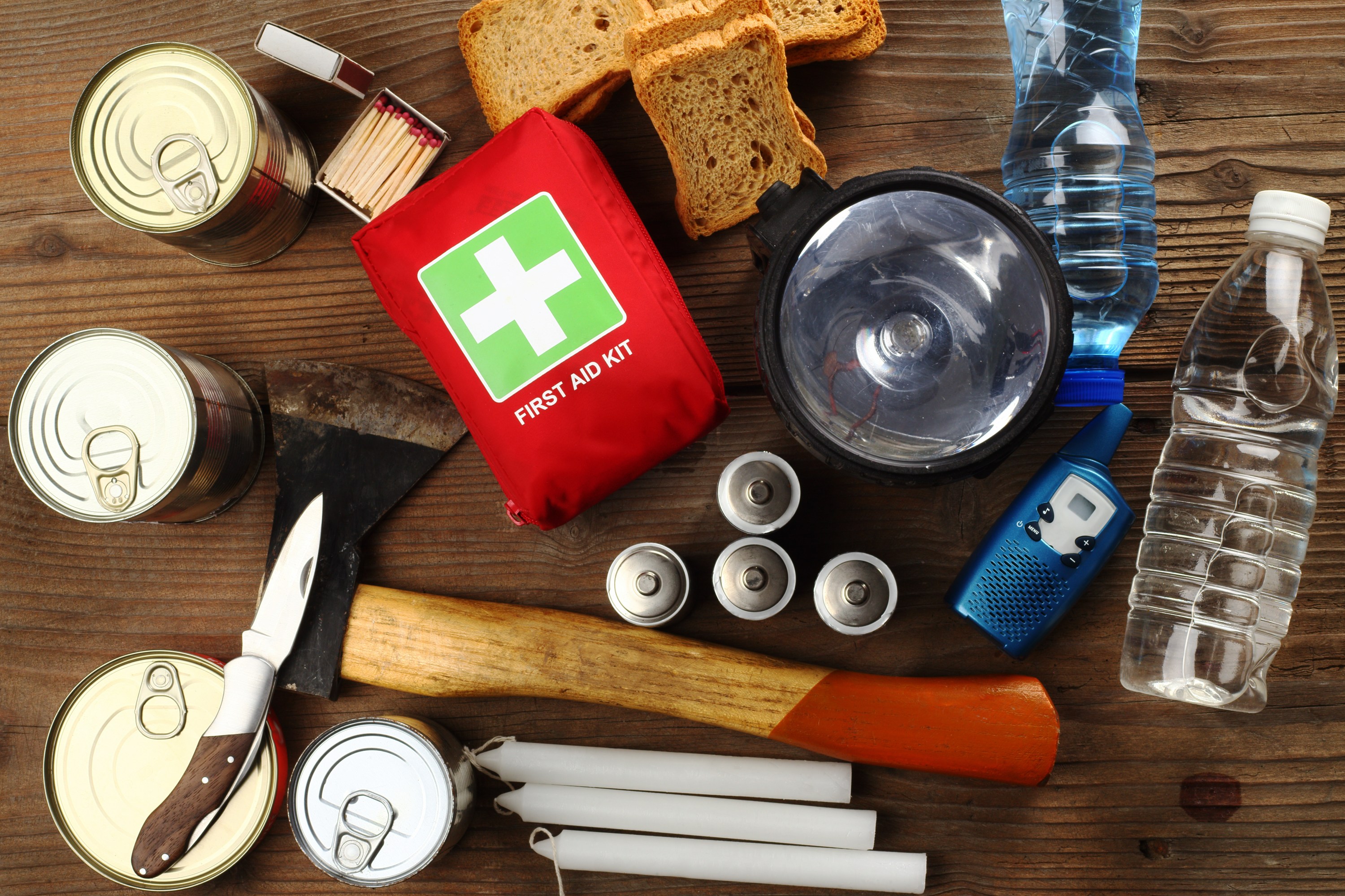 Emergency kit with flashlight, axe, cans, water, radio, bread, and first aid kit on wooden table.