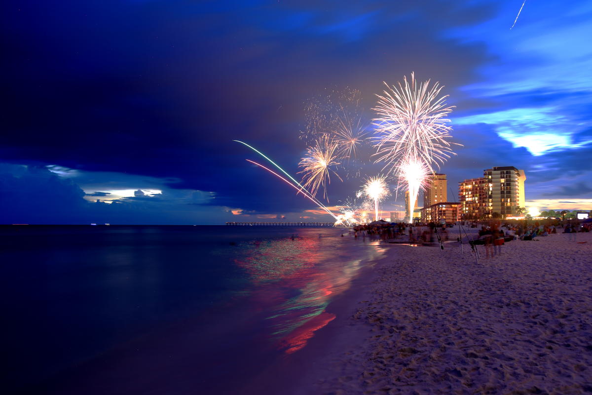 Fireworks over a beach with buildings and a colorful sky at dusk.