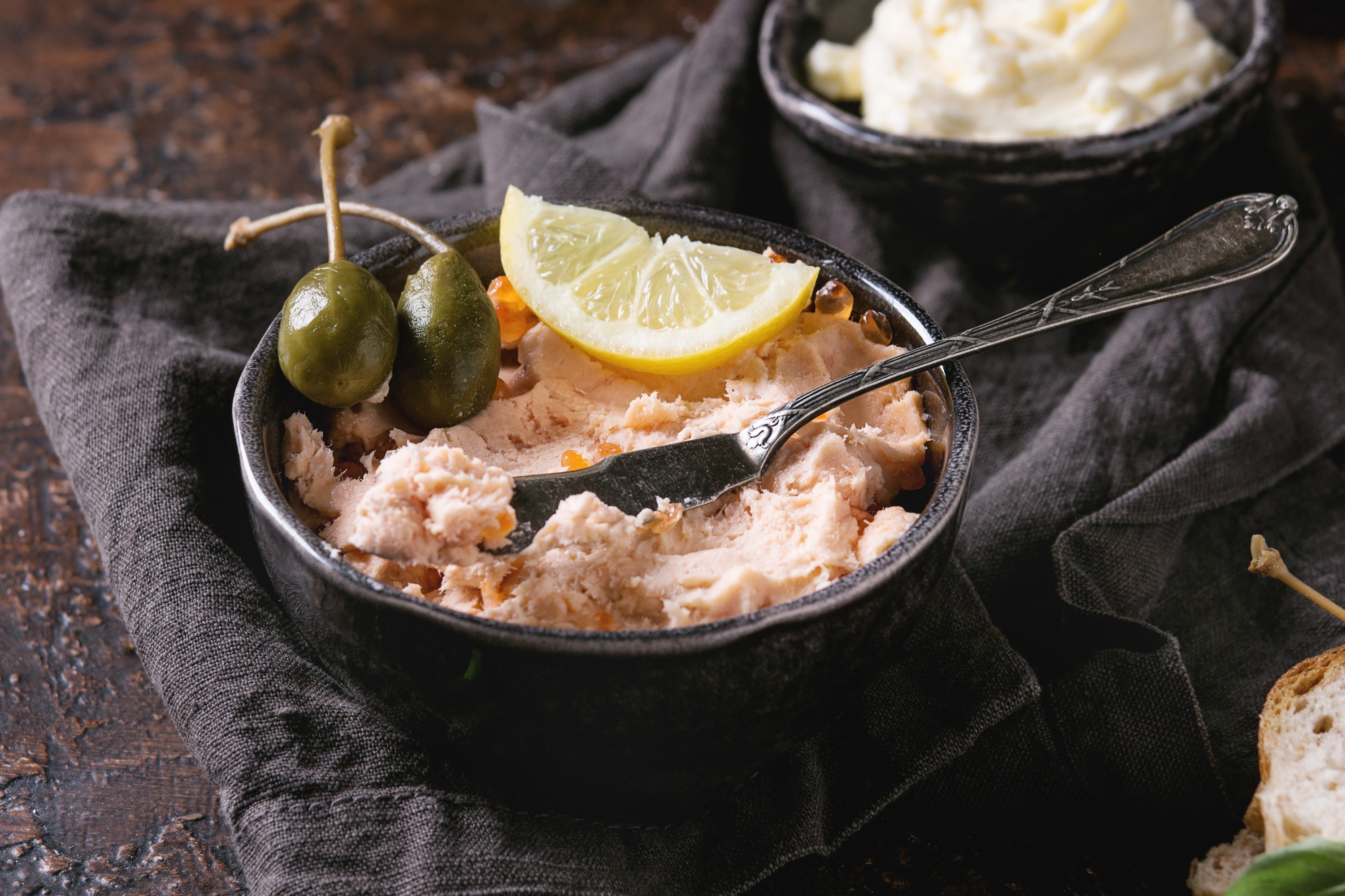Bowl of salmon spread with capers and a lemon slice on a dark cloth.