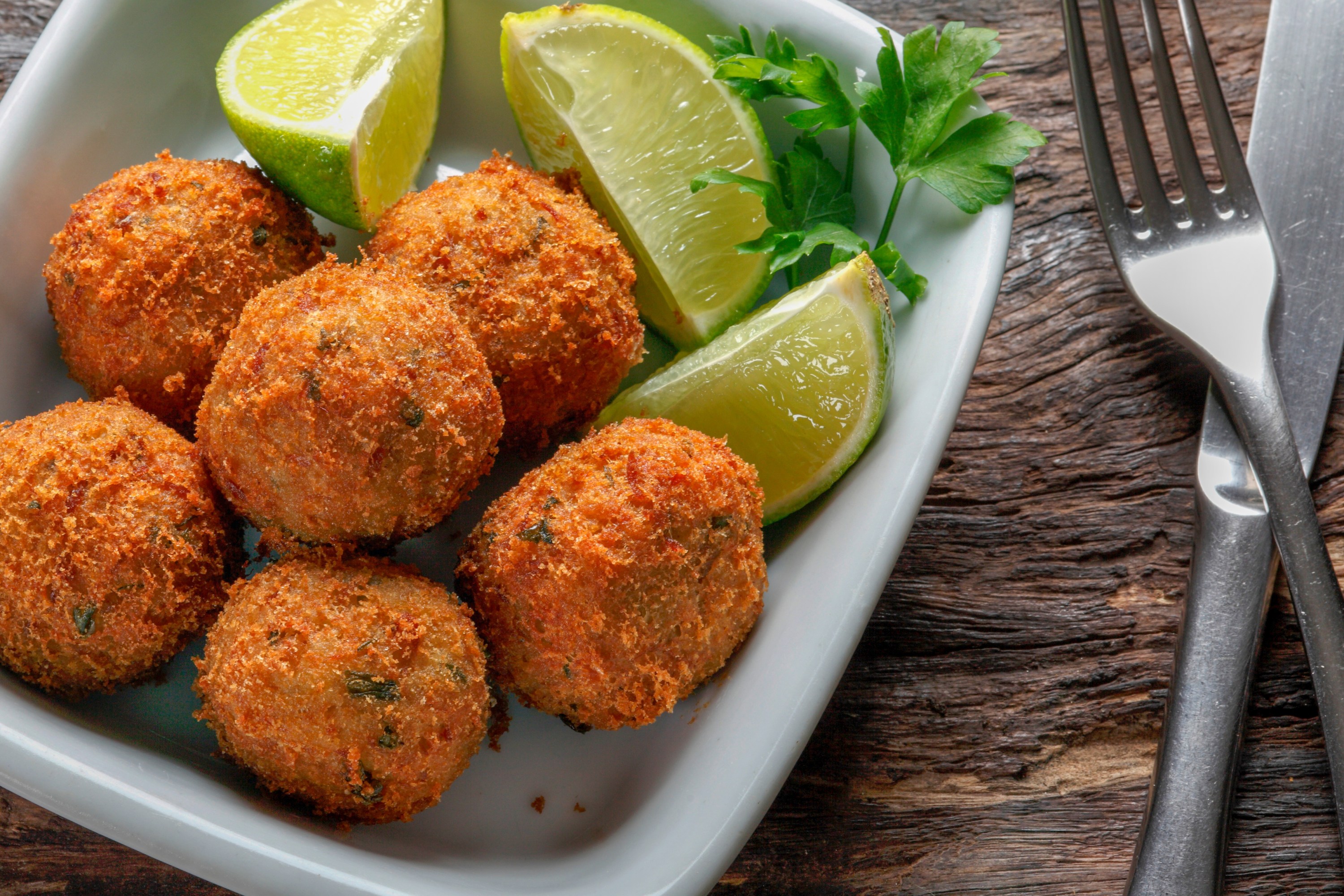 Fried croquettes with lime wedges and parsley in a dish on a wooden table.