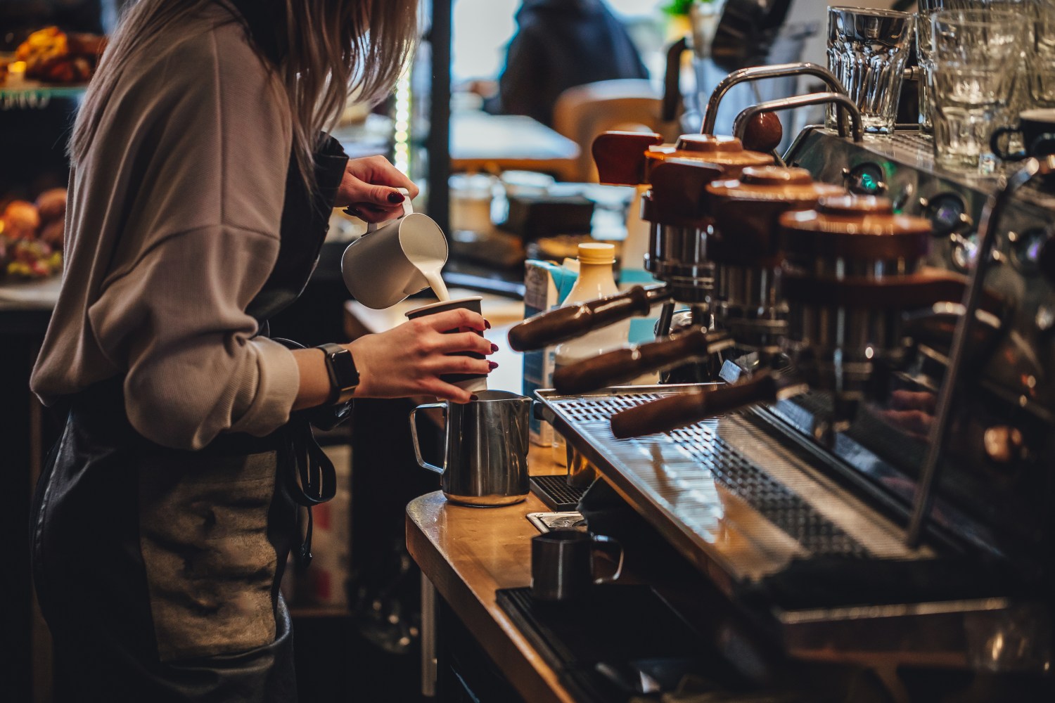 Barista pouring milk into a pitcher next to an espresso machine in a cafe.