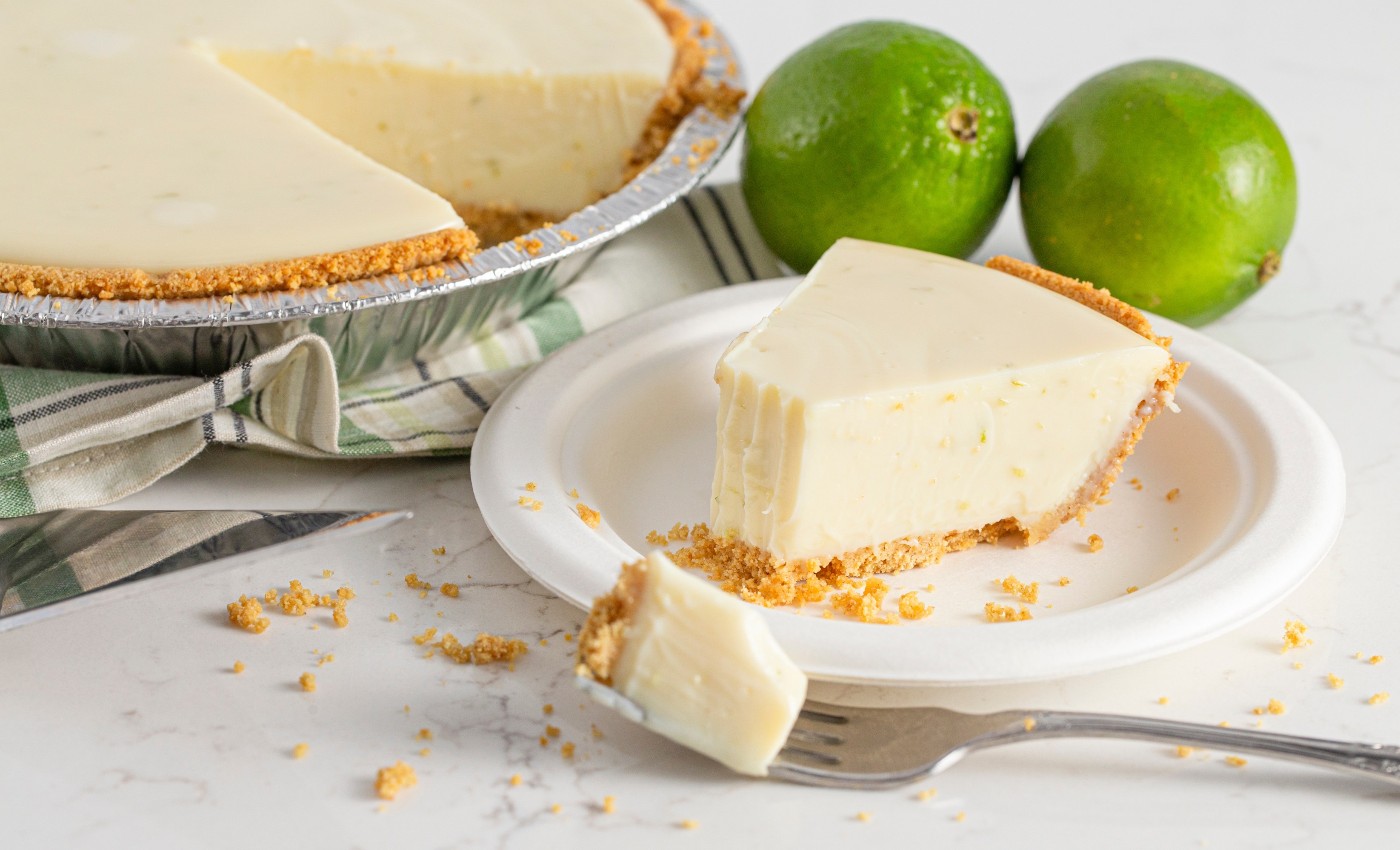 Key lime pie slice on a plate with a fork, whole pie and limes in background.