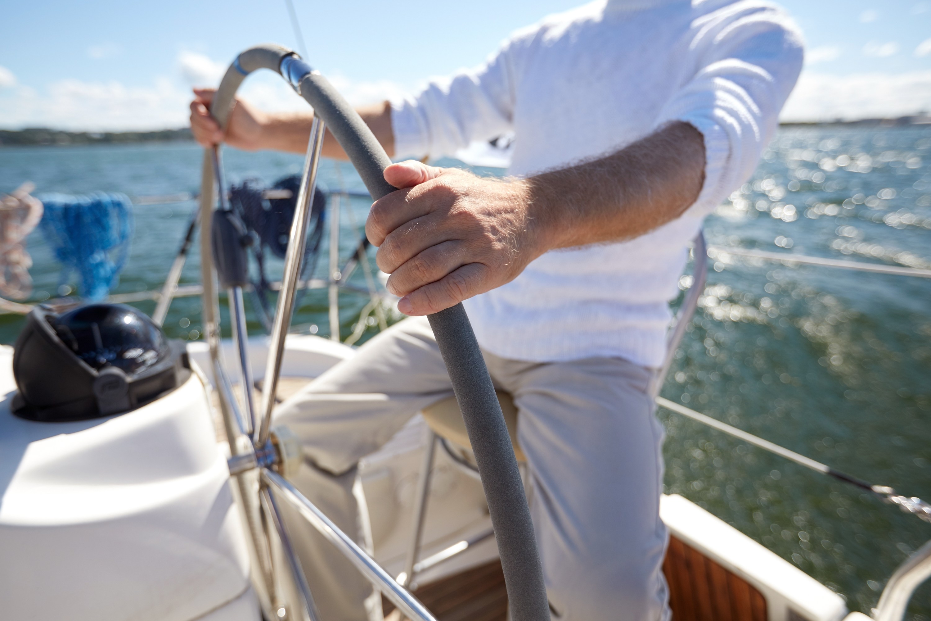 Person steering a sailboat on a sunny day with water in the background.
