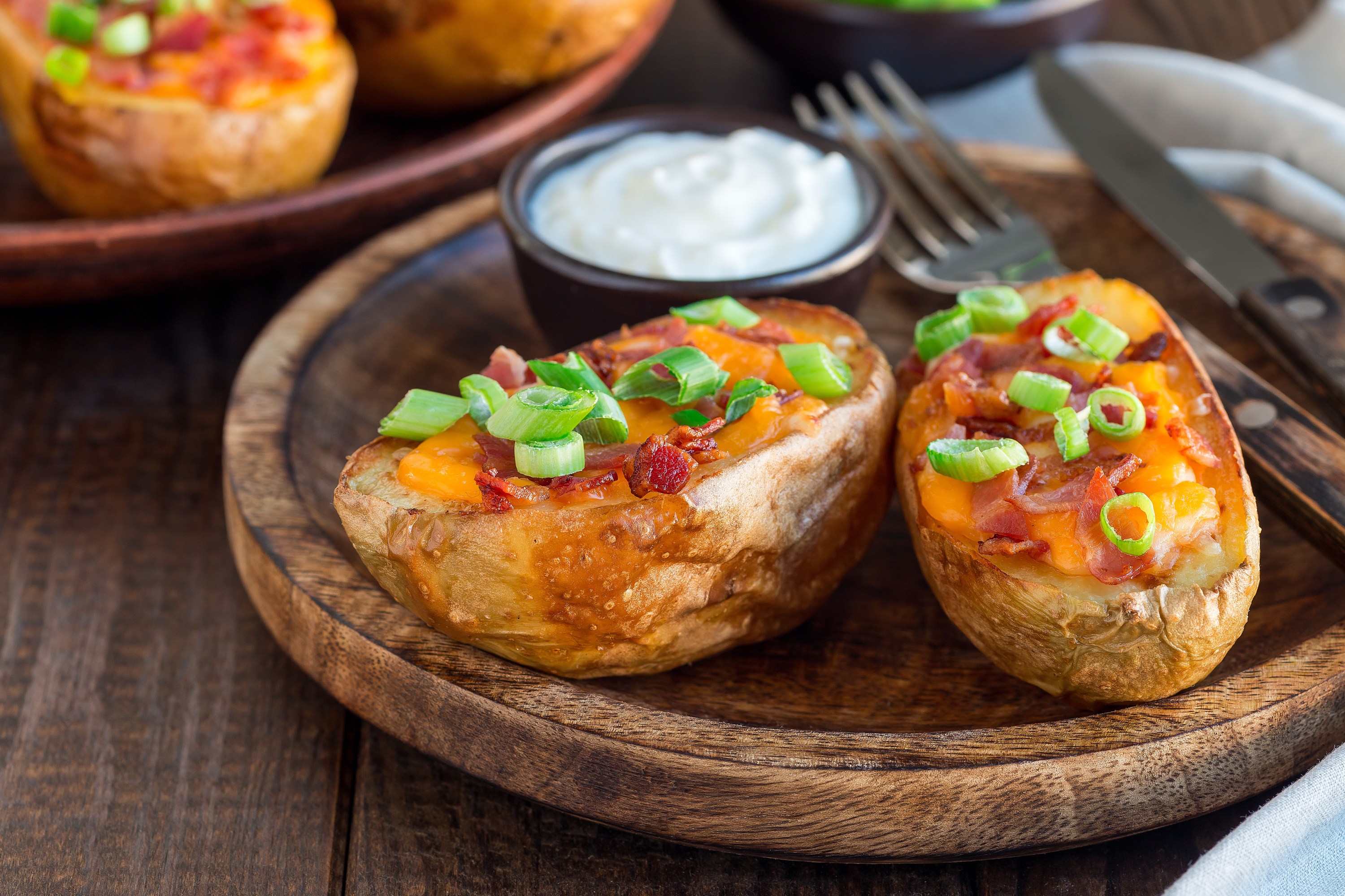 Baked potatoes with cheese, bacon, and green onions on a wooden plate; sour cream in the background.