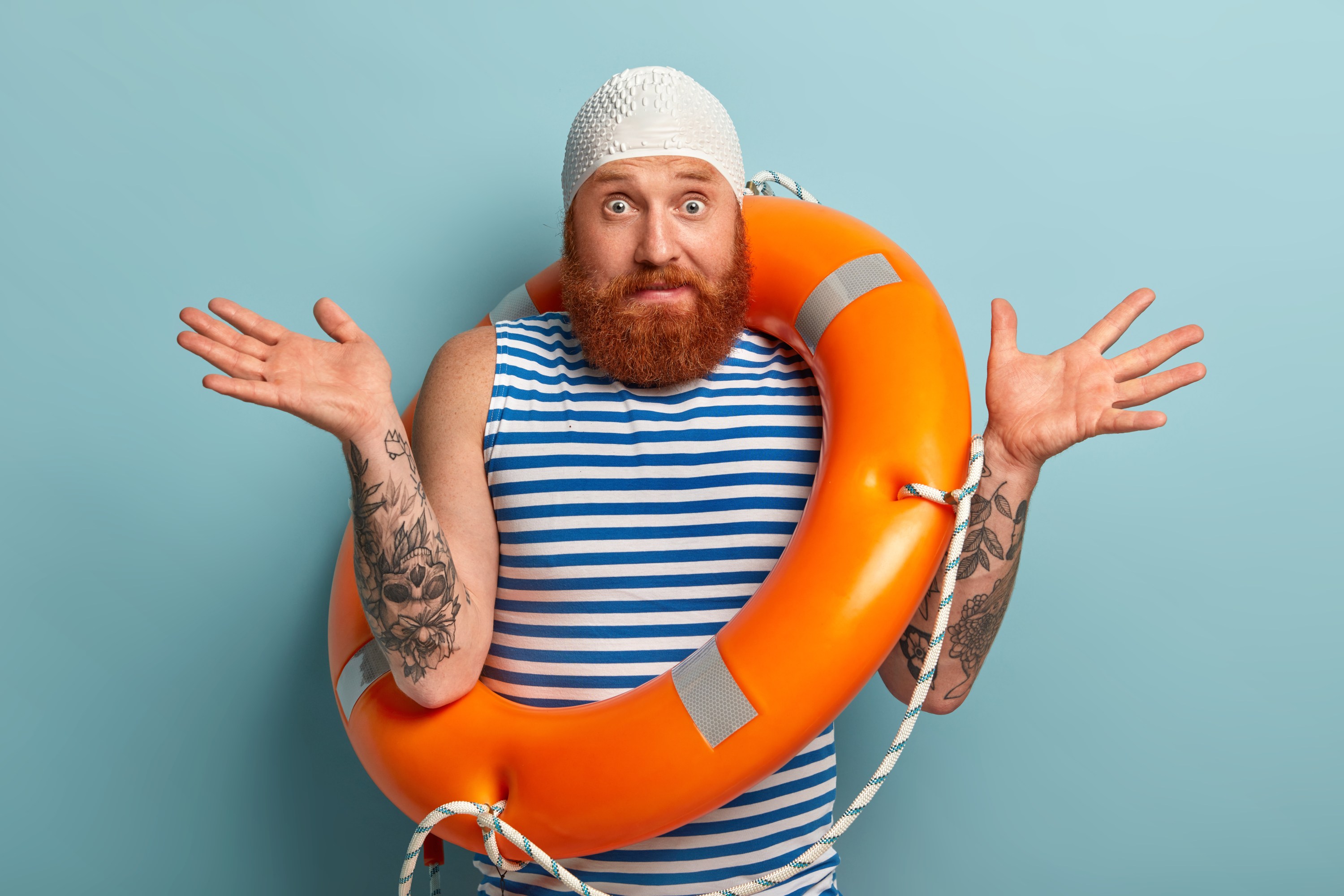 Man with beard and tattoos, in striped shirt and swim cap, wears orange lifebuoy, shrugs with both hands.