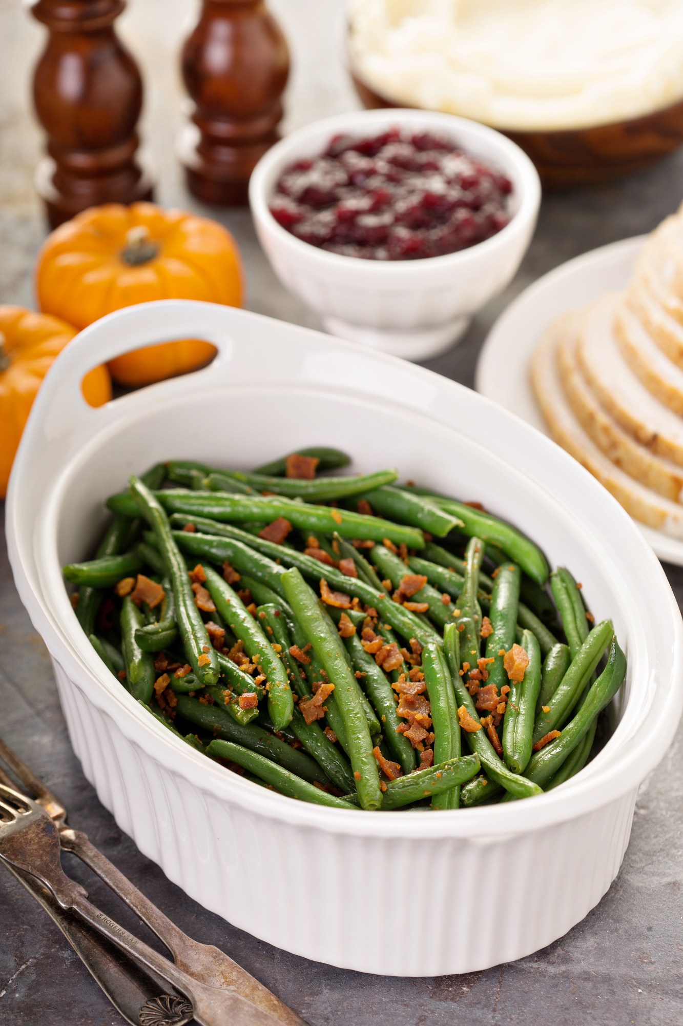White bowl of green beans with bacon bits, surrounded by pumpkins and side dishes.