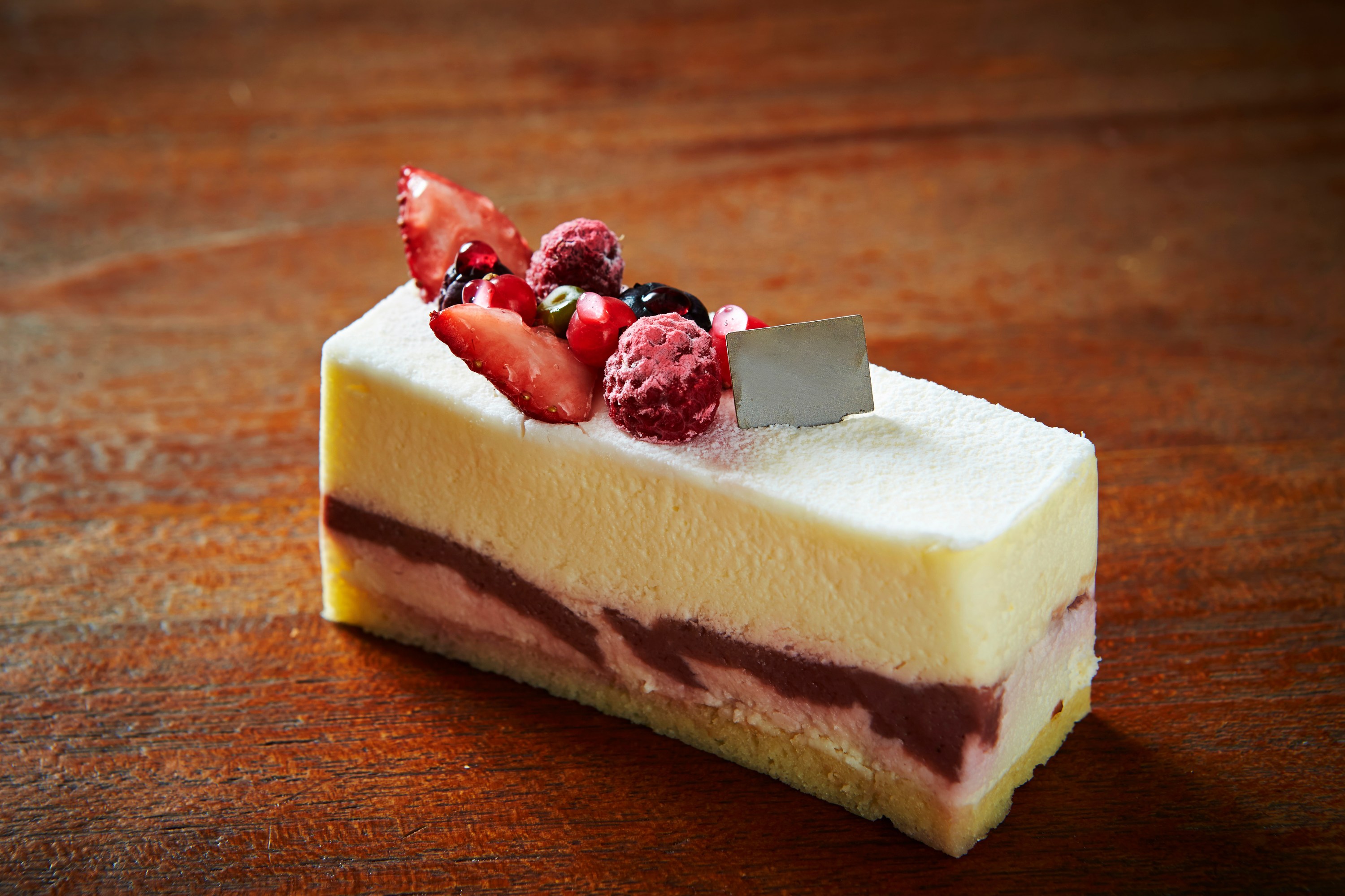 Layered berry cake slice on wooden table, topped with fresh fruit and a small silver decoration.