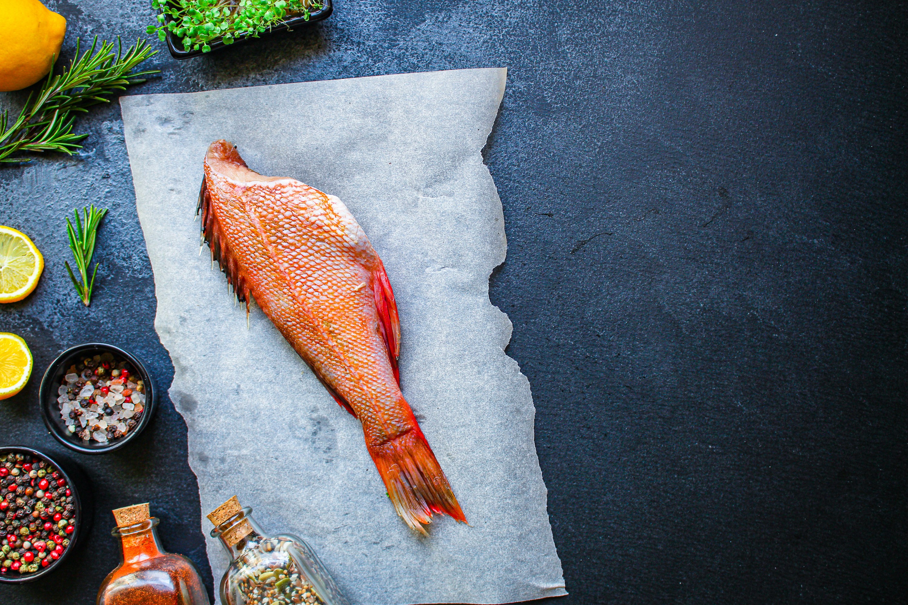 Whole fish on parchment with spices, herbs, and lemon slices on a dark surface.