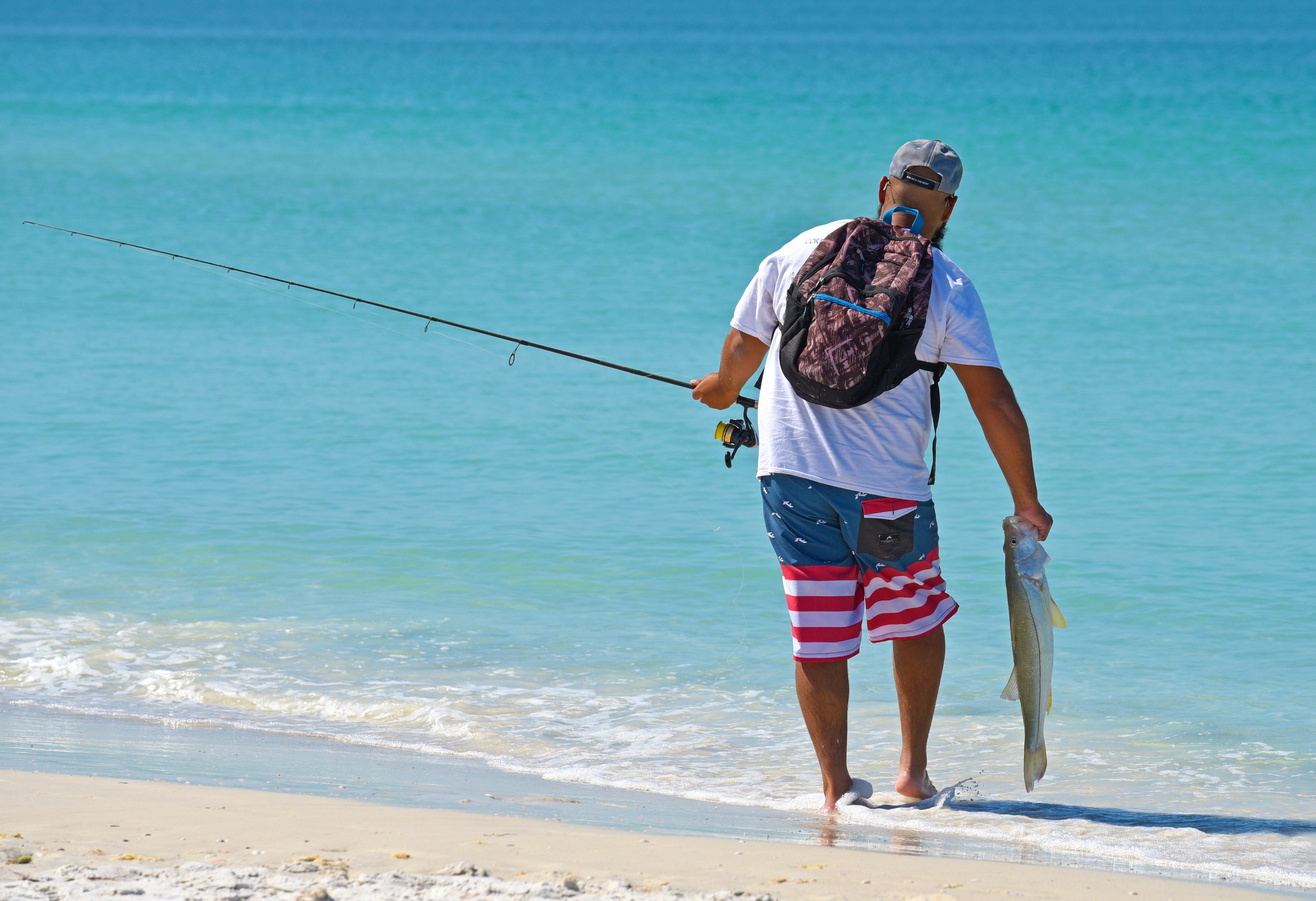 Man in patriotic shorts holding a fish and fishing rod at the beach.