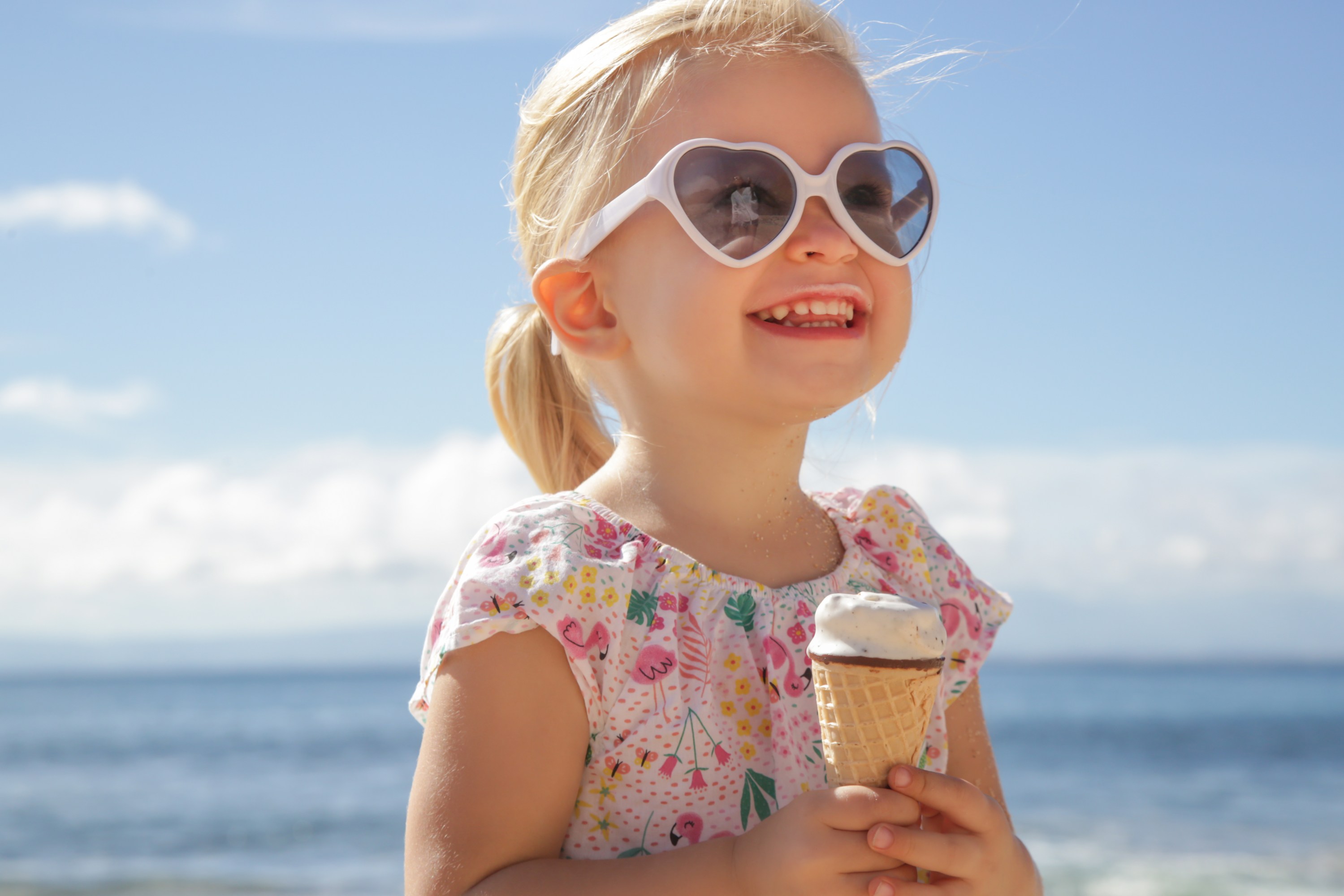 Child in heart-shaped sunglasses holding ice cream at the beach.