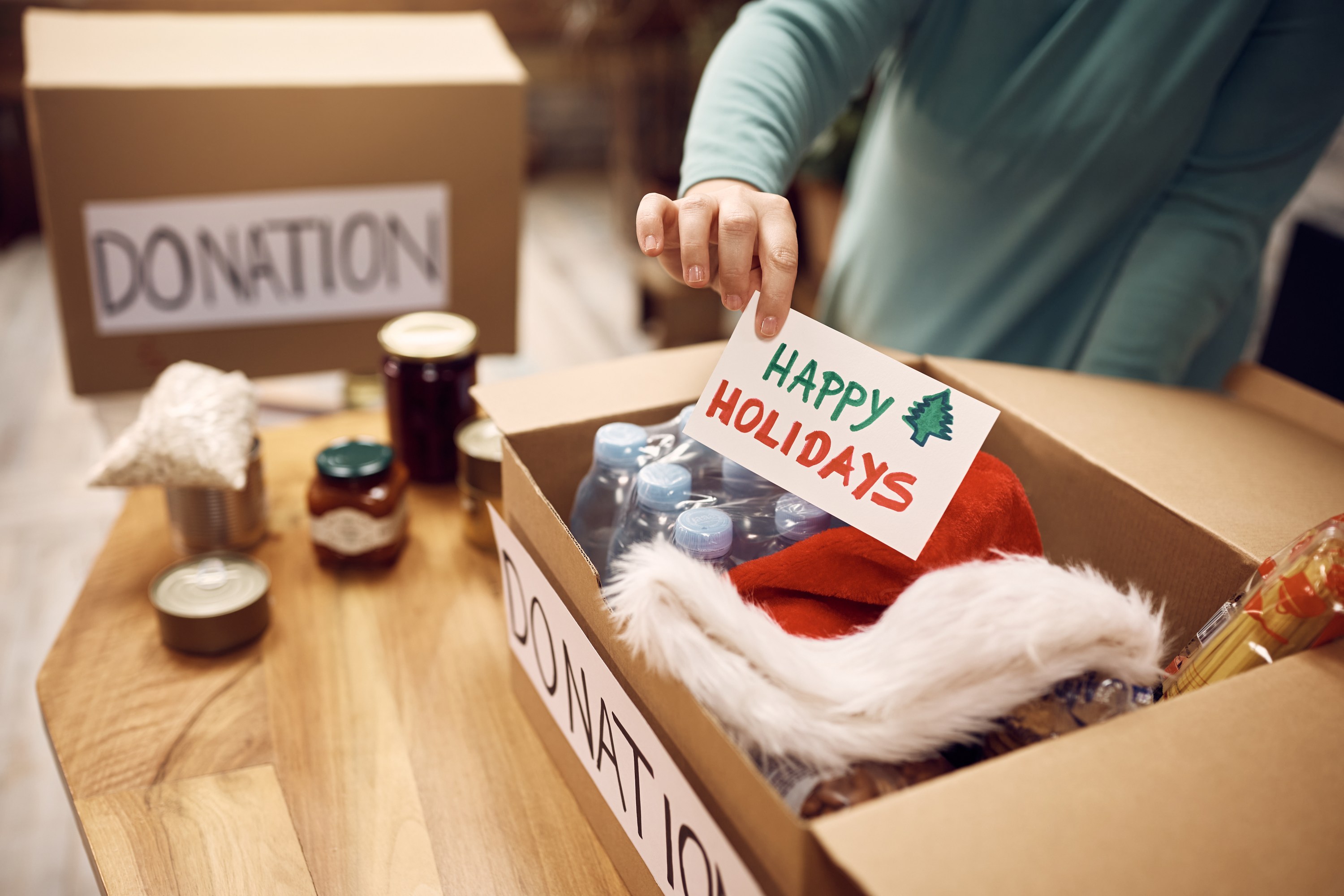 Person placing 'Happy Holidays' card in a donation box with food and a Santa hat.