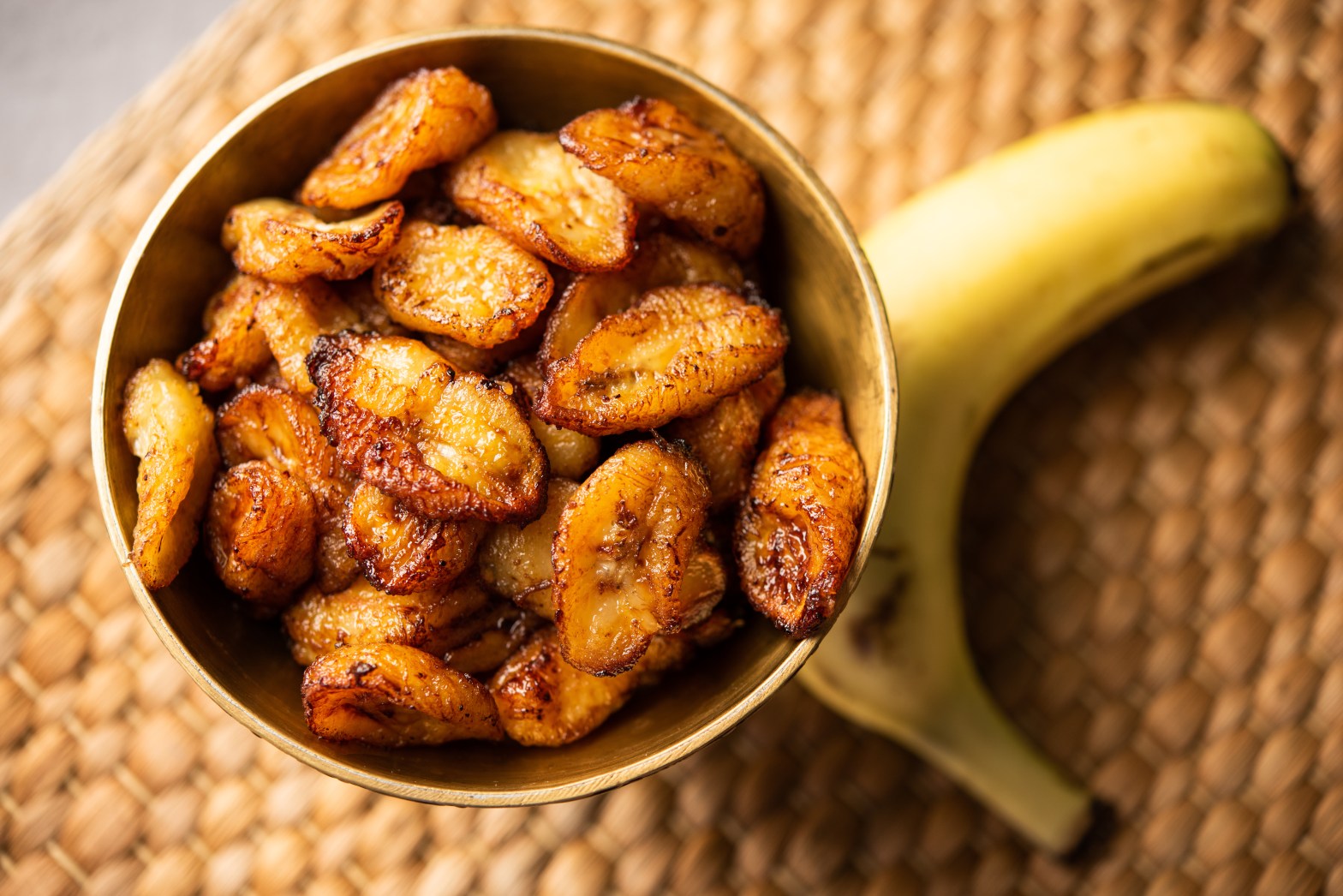 Fried plantains in a bowl next to a ripe banana on a woven mat.