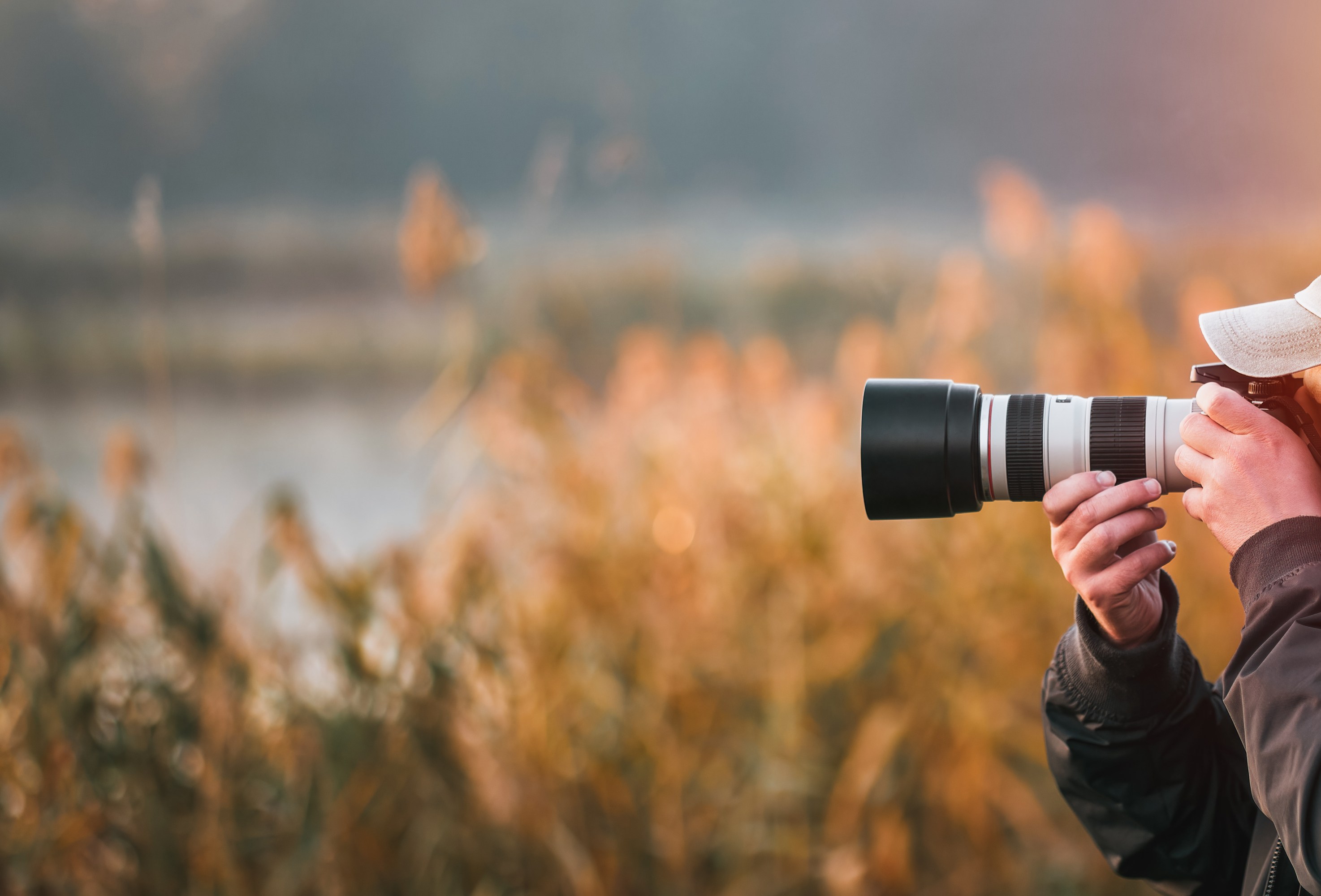 Person with a hat using a camera with a large lens in a field at sunset.
