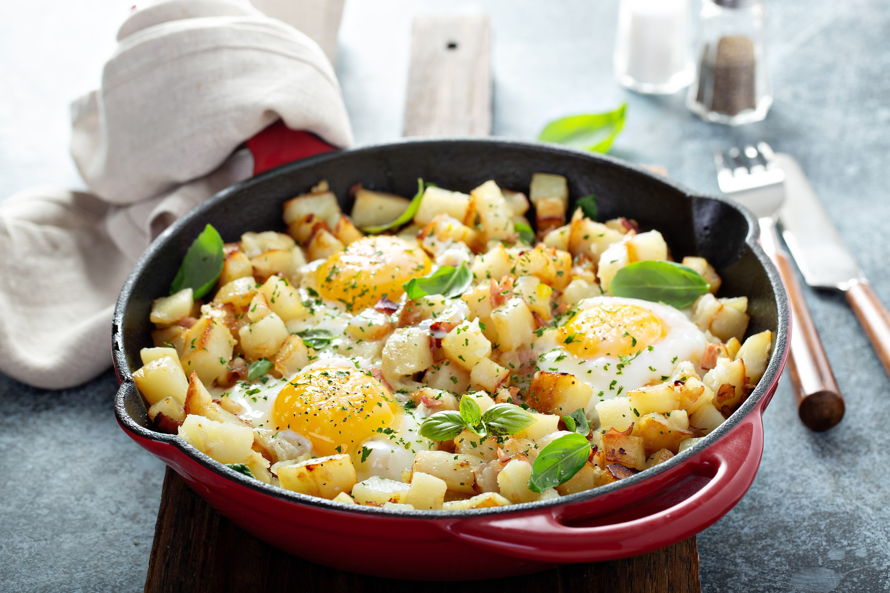 Fried eggs with diced potatoes and herbs in a red skillet on a gray surface.