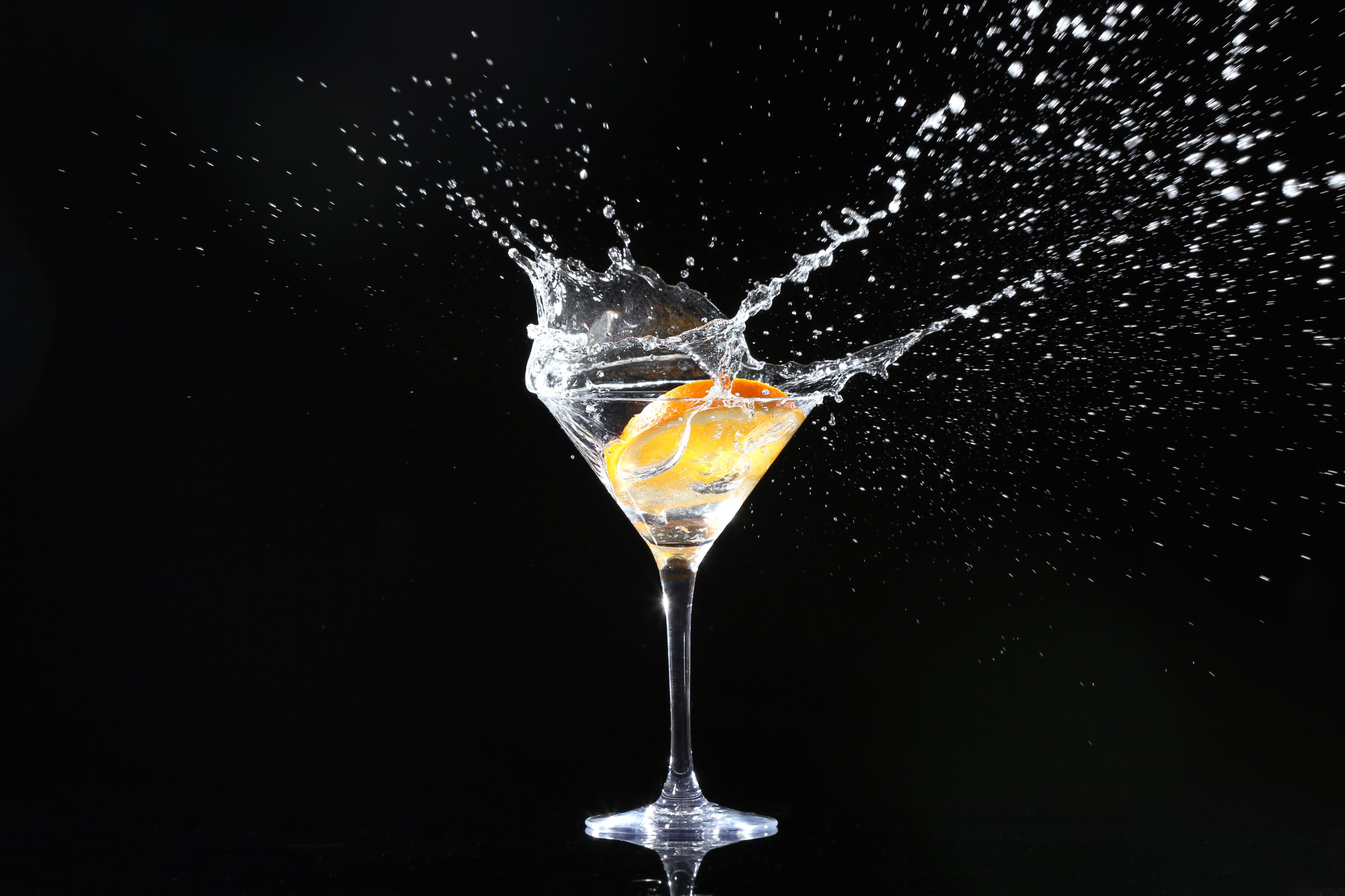 Martini glass with liquid splashing out and orange twist on black background.