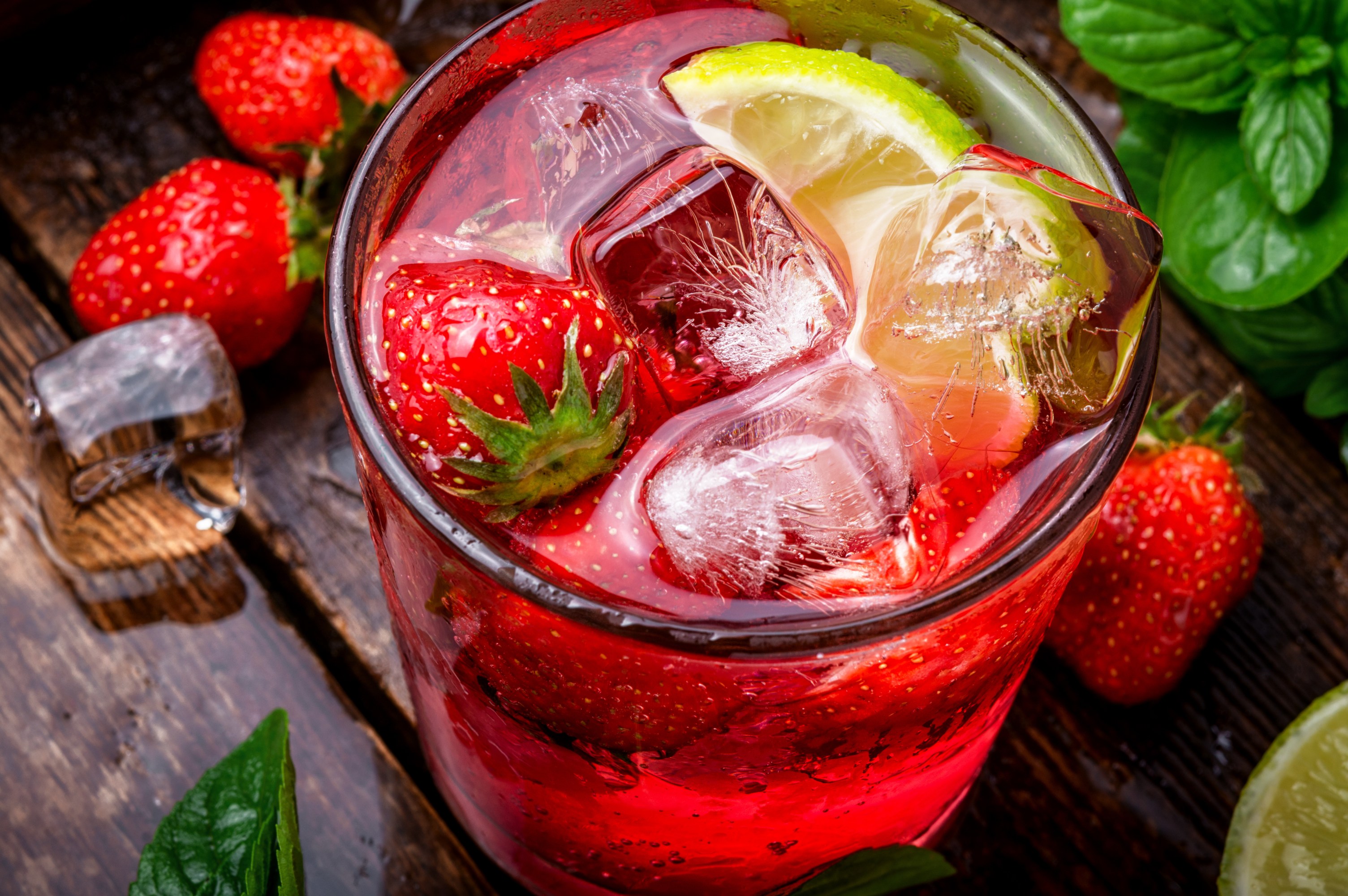 Glass of red drink with strawberries, lime slices, and ice cubes on wooden table.