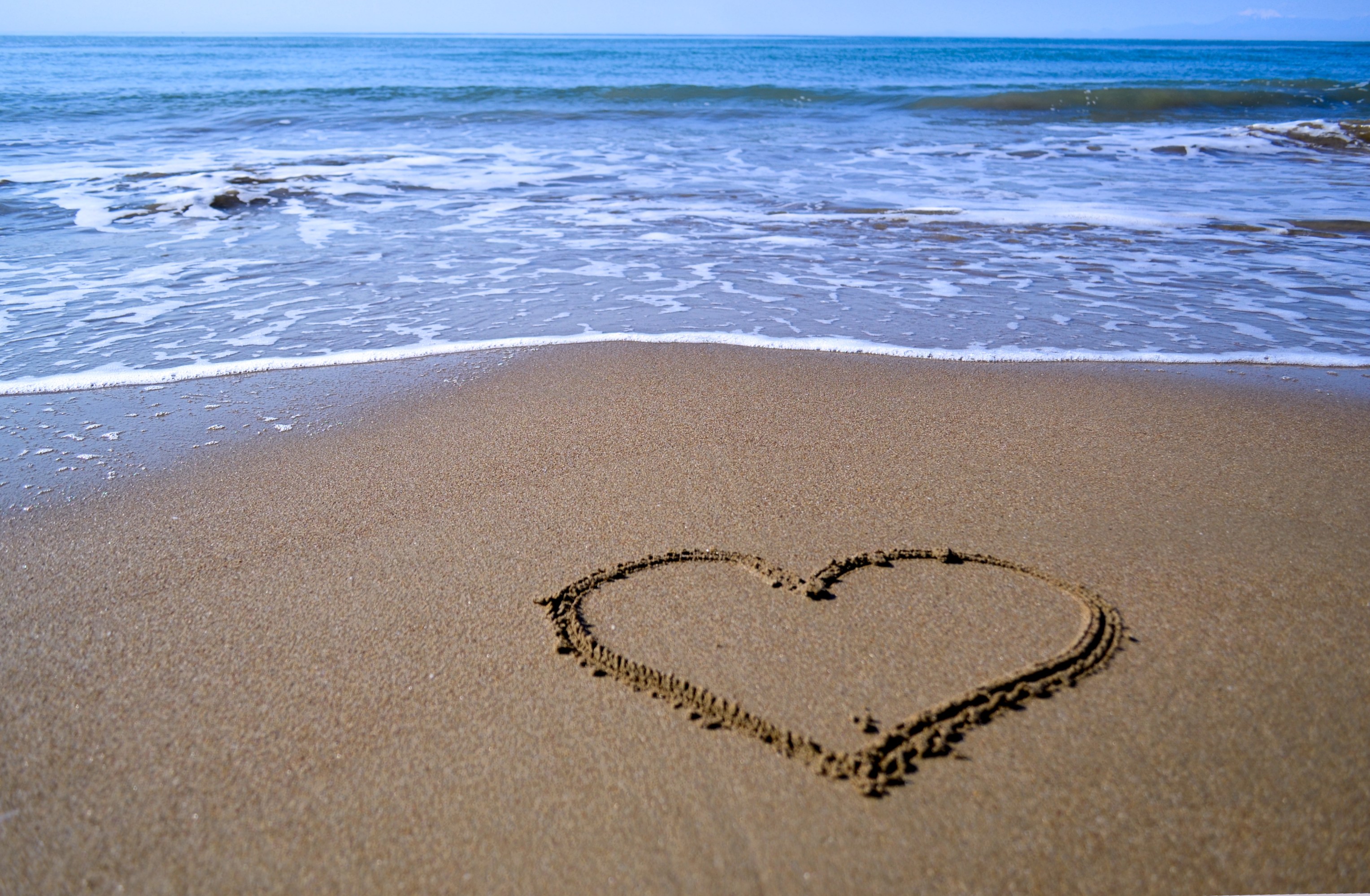 Heart drawn in sand on beach with ocean waves in background.