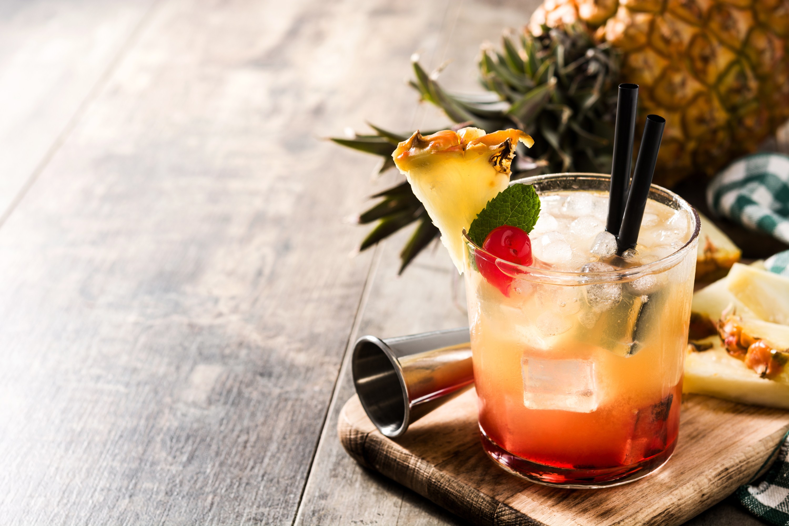 Tropical cocktail with pineapple slice and cherry on wooden board, pineapple in background.