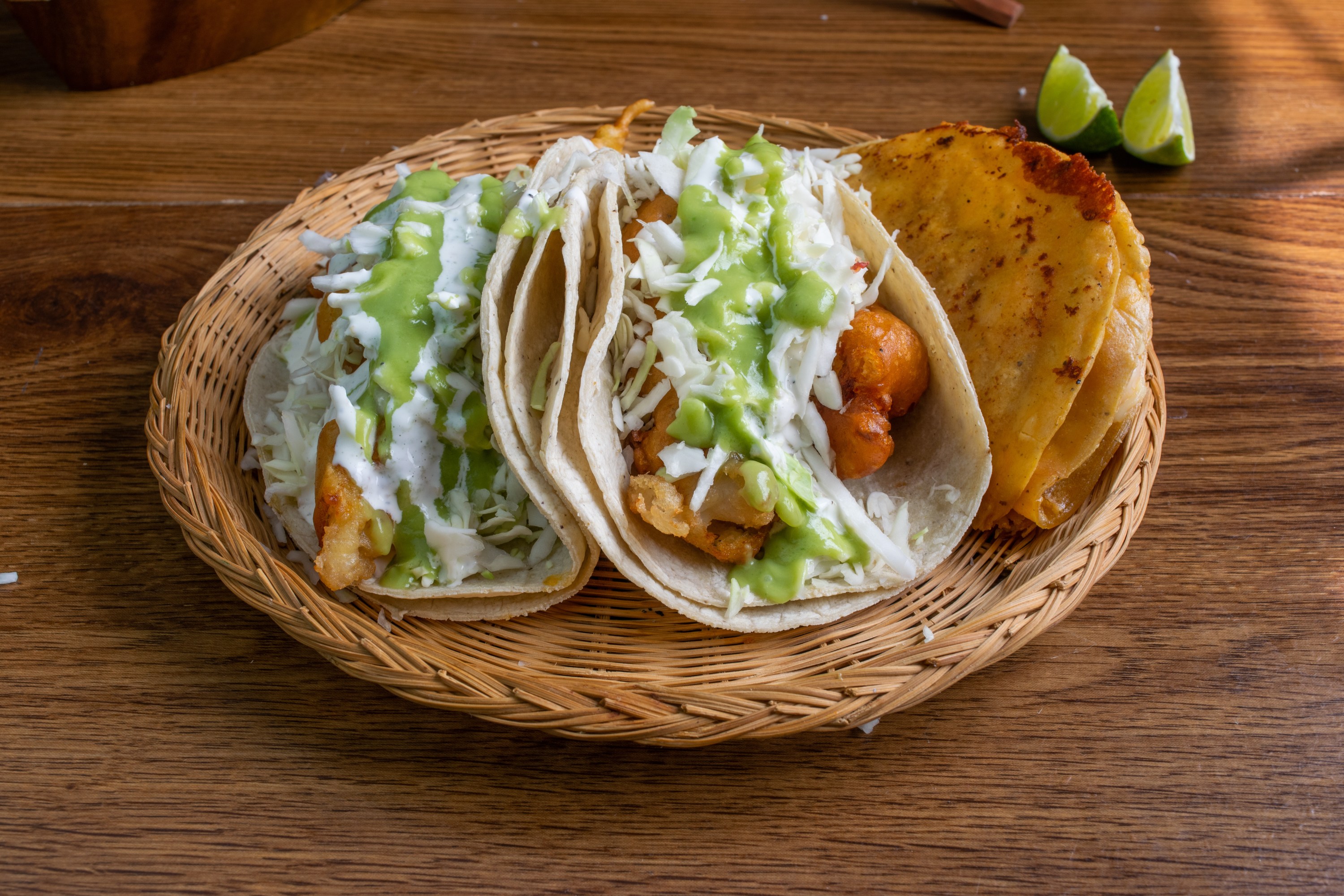 Basket with two tacos topped with green and white sauces, next to folded tortillas, on a wooden table.