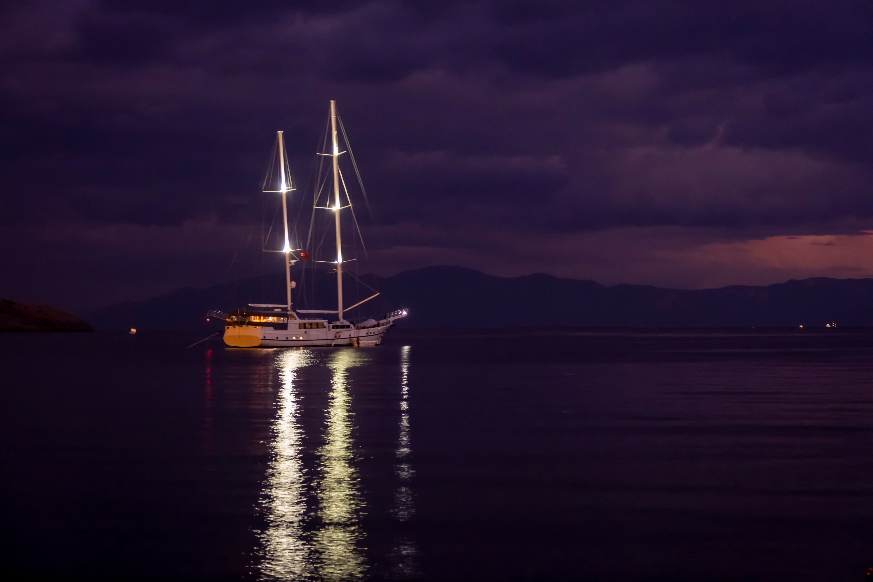 Sailboat with lights on, reflecting on calm water at dusk, under a dark, cloudy sky.