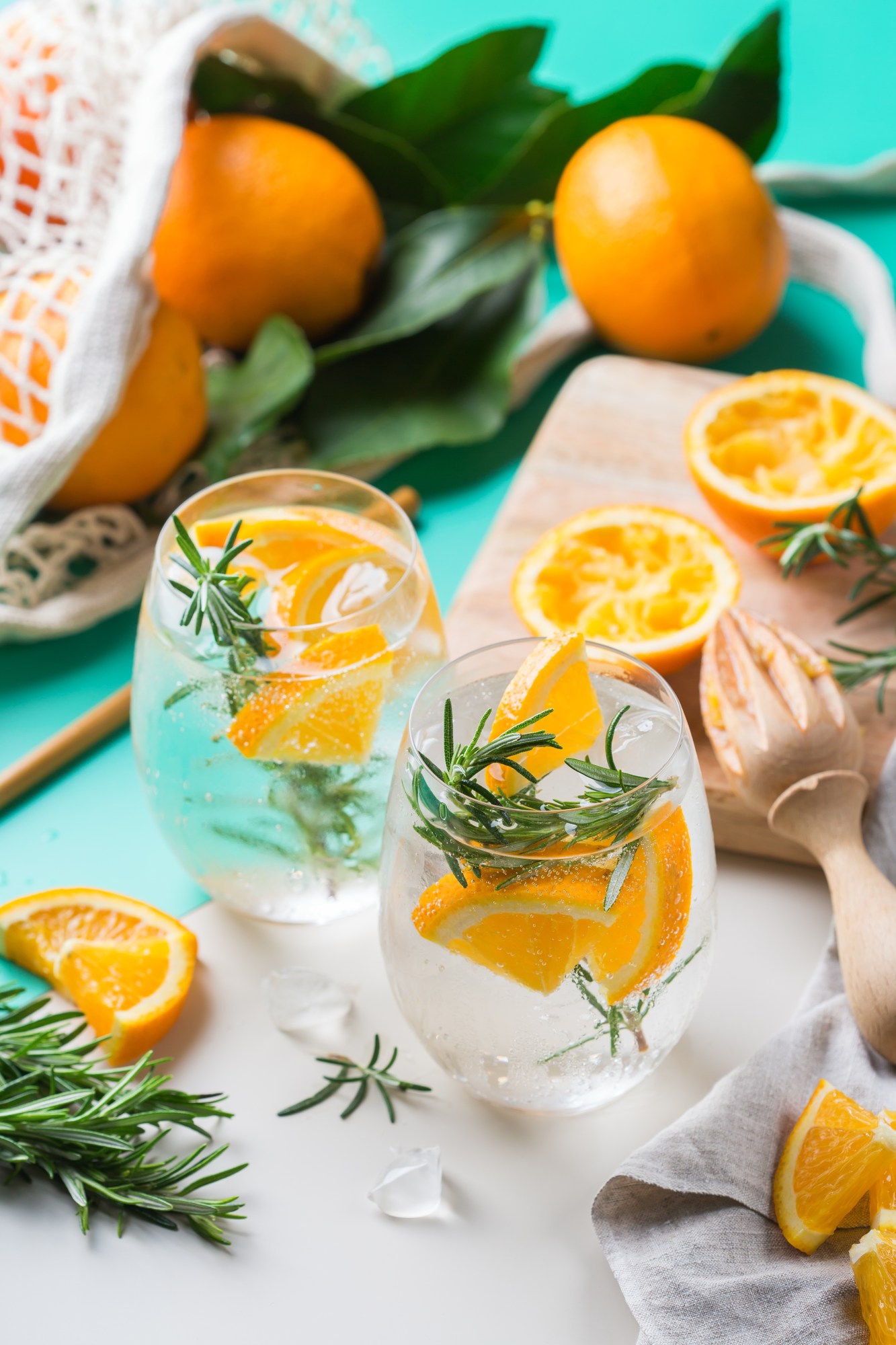 Two drinks with orange slices and rosemary on a table with whole oranges.