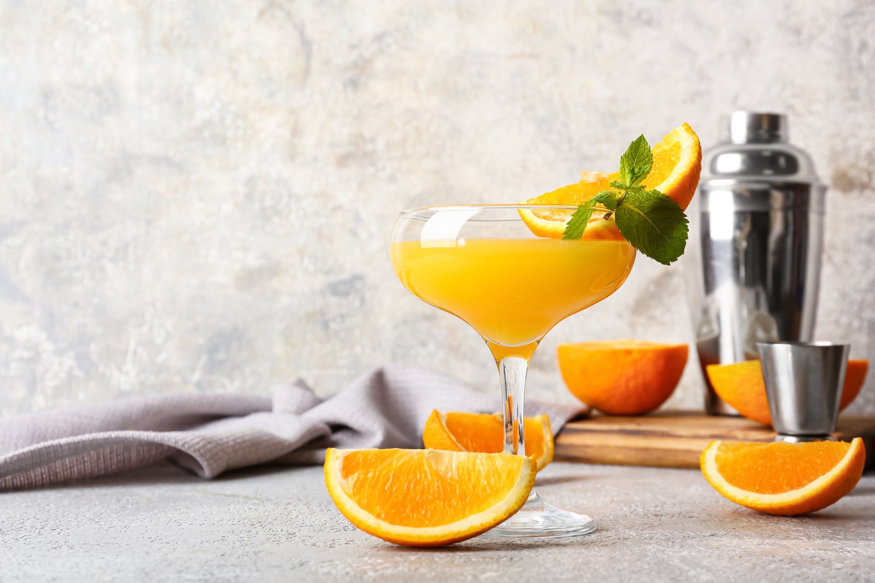 Orange cocktail with mint garnish in a glass, surrounded by orange slices and a shaker.