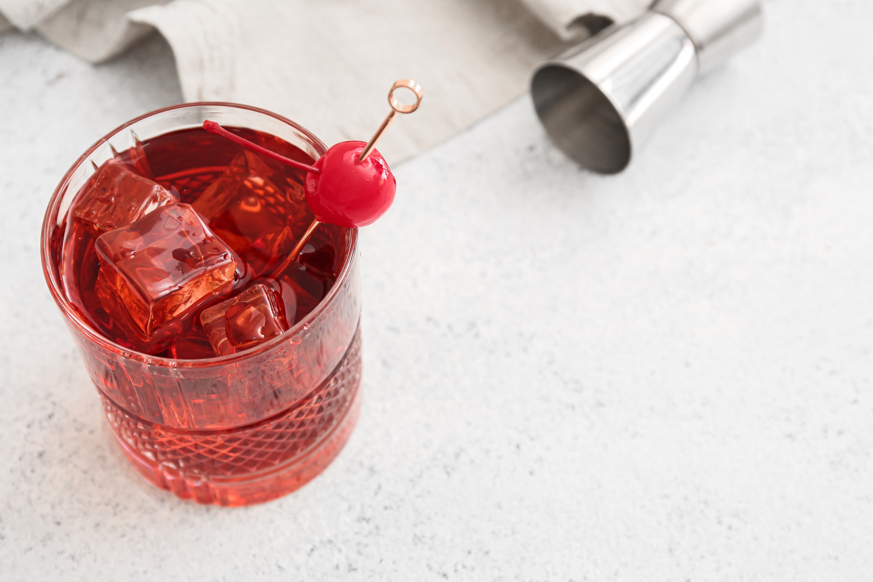 Red cocktail with ice and a cherry garnish in a glass on a light surface.