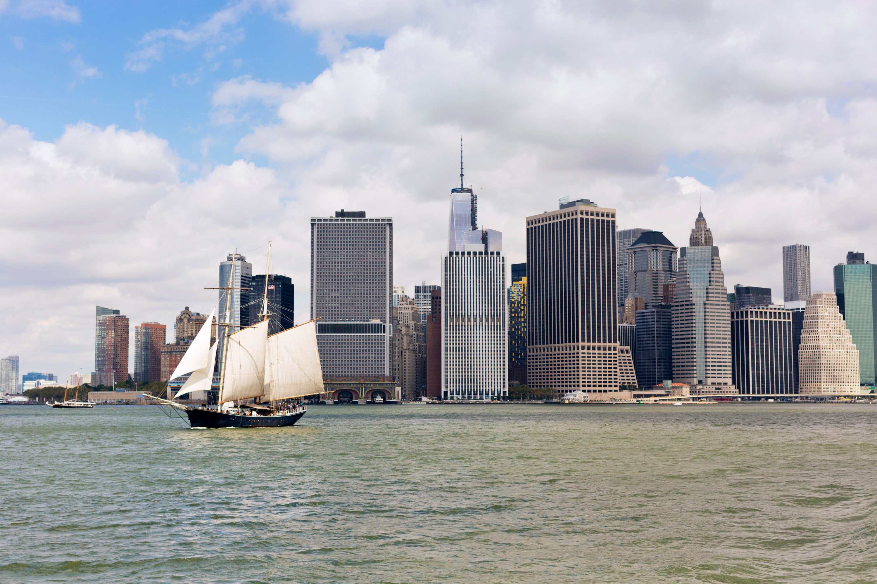 Sailboat on water in front of city skyline with tall buildings under a cloudy sky.