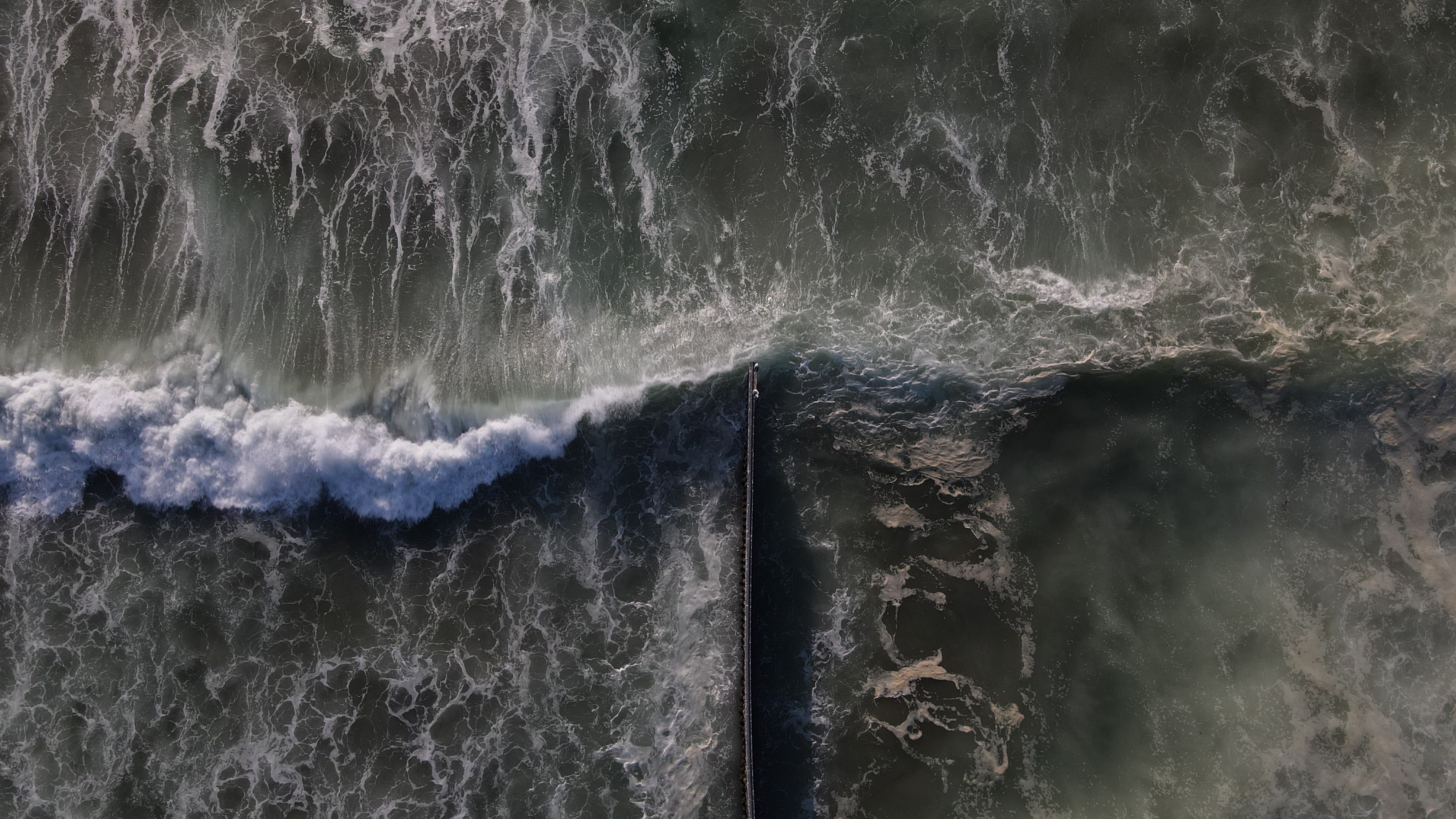 Aerial view of waves crashing against a pier extending into the ocean.