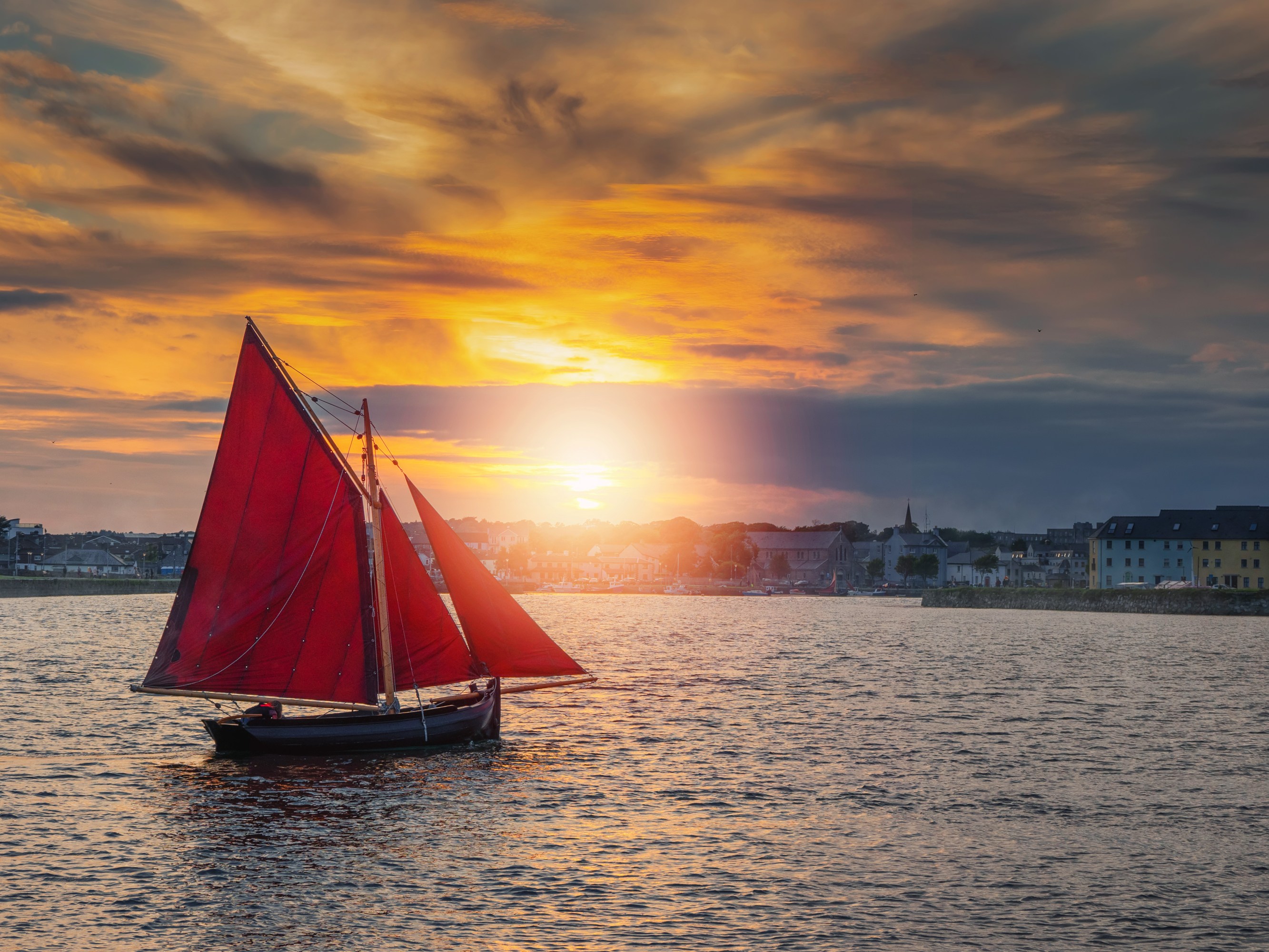Sailboat with red sails on water at sunset with town skyline in background.
