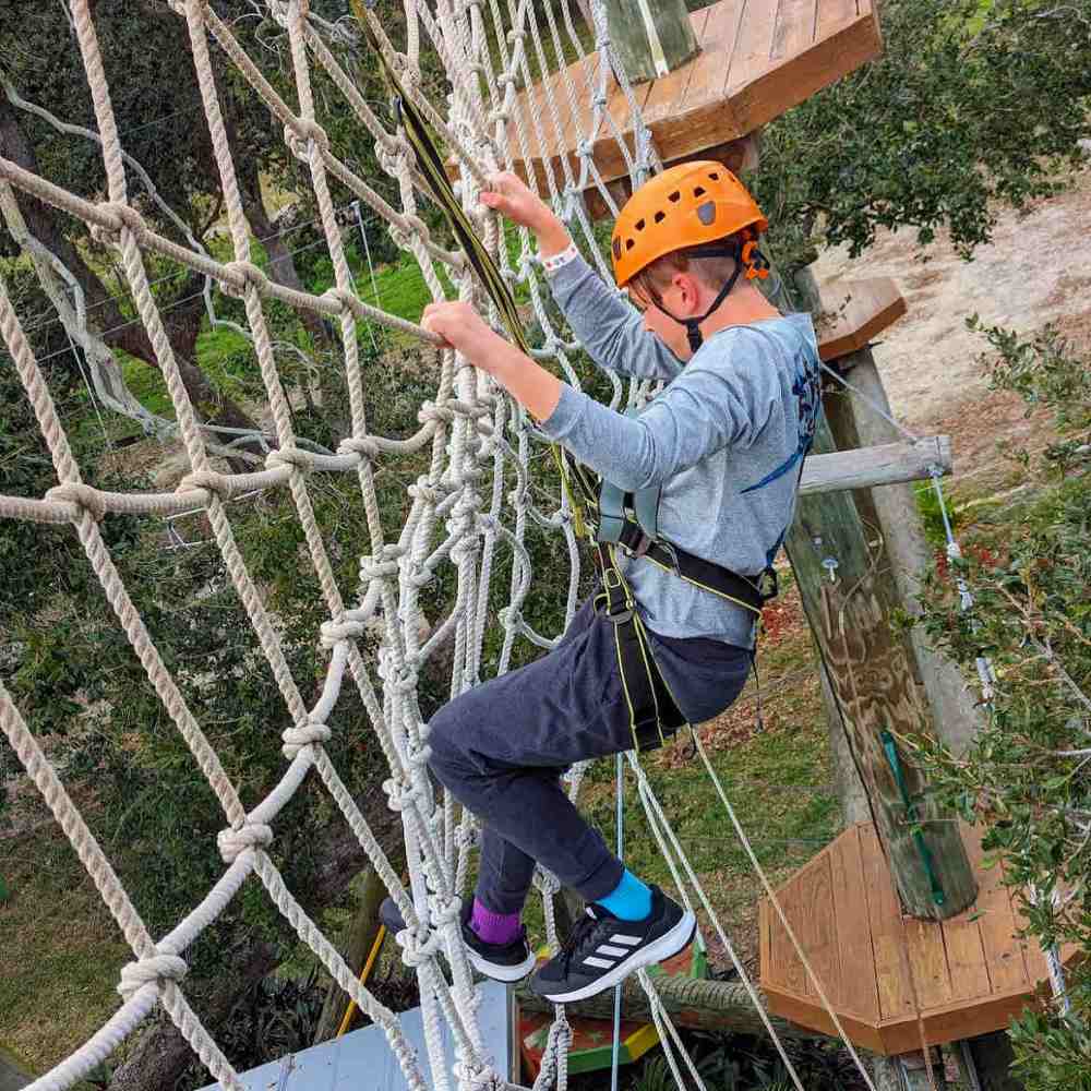 Person in helmet and harness climbing a rope net course outdoors.